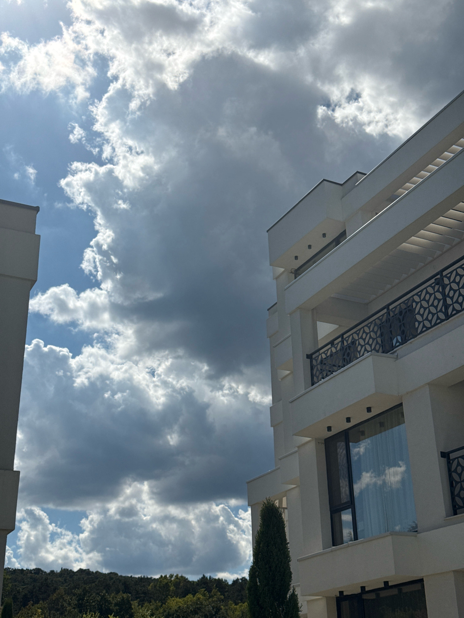 The image shows a modern white apartment building with balconies and large windows, set against a dramatic sky filled with both bright white and dark gray clouds. The building's architecture is characterized by clean lines and geometric shapes. In the foreground, a tall, slender evergreen tree stands next to the building. The background reveals a densely wooded hillside, suggesting a natural setting surrounding the urban structure. The scene is likely located in Sozopol, Bulgaria, given the building's style which is common in newer developments in coastal towns. There are no people visible in the image.