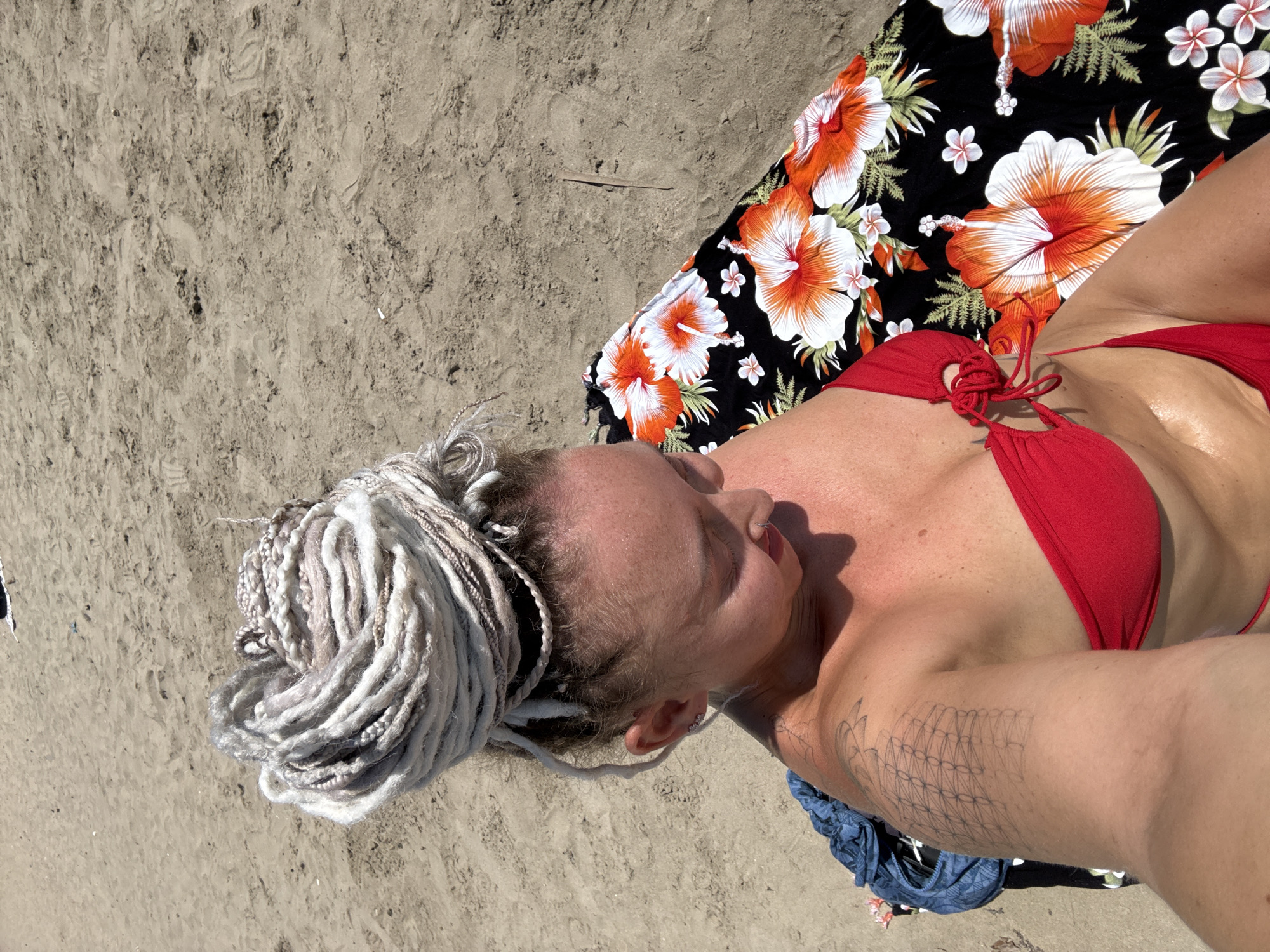 A woman with blonde dreadlocks piled on her head sits on a beach towel with a floral pattern of orange and white hibiscus flowers on a sandy beach. She is wearing a red bikini top. The sand is light brown and shows some footprints.
