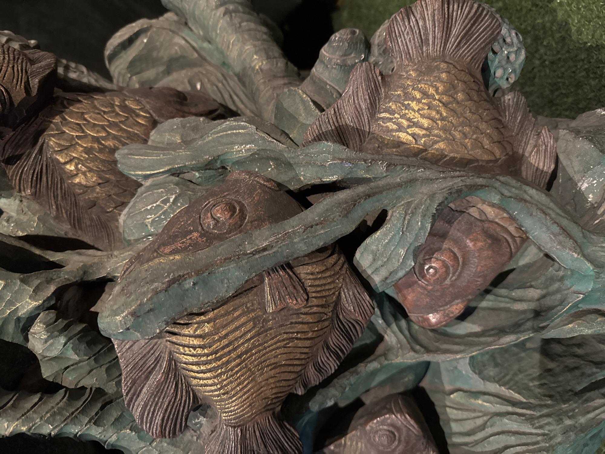 The image shows a close-up, detailed view of a bronze sculpture featuring multiple fish intertwined with stylized aquatic plants. The sculpture exhibits a patina, with some areas appearing bronzed or golden, while others are coated in a greenish-blue verdigris. The fish are intricately detailed with visible scales and fins, suggesting a dynamic underwater scene. The texture of the sculpture is rough and carved, with pronounced lines and edges on both the fish and the plant elements. The background is dark and out of focus, with a hint of green, possibly grass or moss, in the lower right corner. There are no people or actions depicted; it is a static object. The scene is not identifiable as being in Kuta, Indonesia, or any specific city.