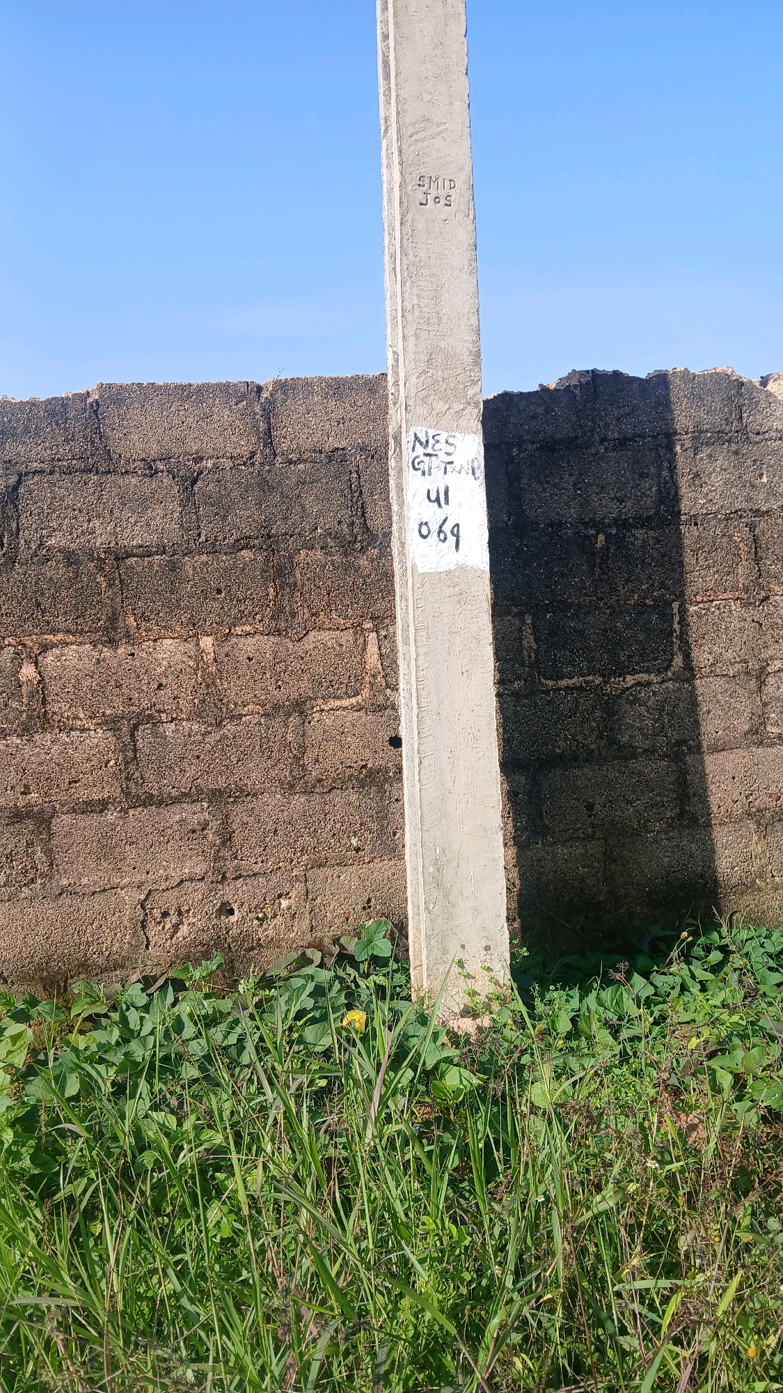 The image depicts a concrete pole in front of a brick wall, located in Gura Top, Nigeria. The pole has markings on it, including "SMID Jos" carved into the upper section and "NES GTTA 41 069" written in black ink on a white patch lower down. The brick wall appears to be old and weathered, with some sections crumbling. Lush green vegetation, including tall grass and leafy plants, grows at the base of the wall and around the pole. The sky is clear and blue, indicating a sunny day. The scene is static, with no people or animals present. The overall impression is one of an outdoor, possibly rural or semi-urban environment, with evidence of infrastructure and natural growth.