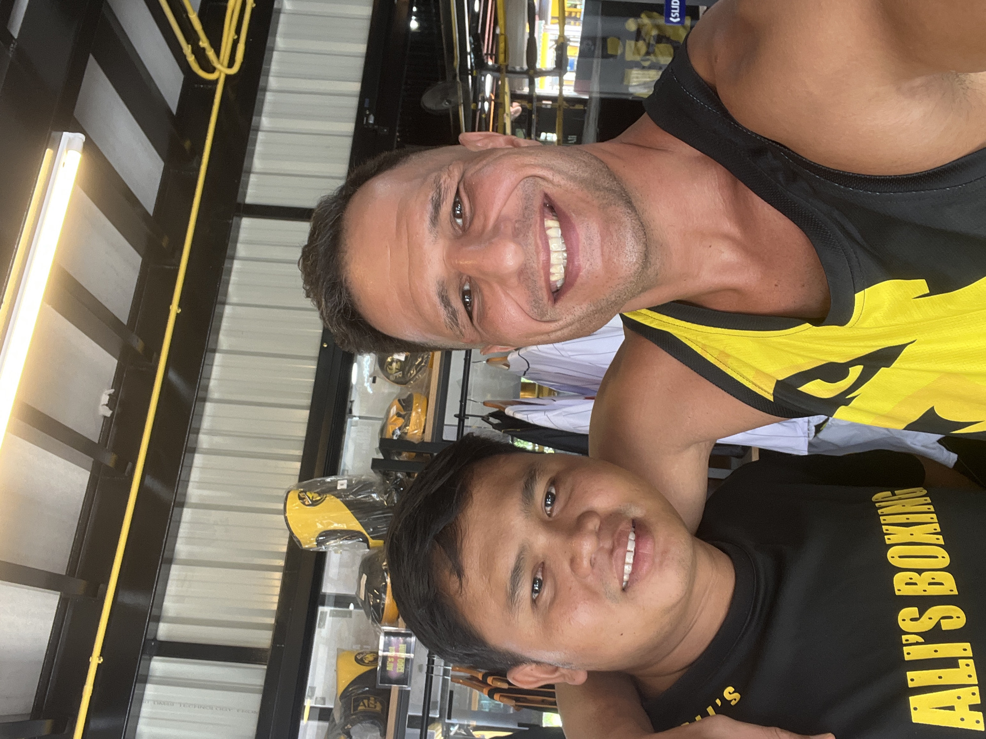 Two men are posing for a selfie in what appears to be a boxing gym in Chalong, Thailand. The man on the left, with short dark hair, is wearing a black t-shirt with yellow lettering that reads "ALI'S BOXING". The man on the right, with short dark hair and a muscular build, is wearing a black and yellow tank top. Both men are smiling. In the background, there are shelves stocked with boxing equipment, including headgear and gloves, all predominantly in yellow and black. There is also a boxing ring visible in the background. The environment suggests a commercial establishment focused on boxing training and merchandise.