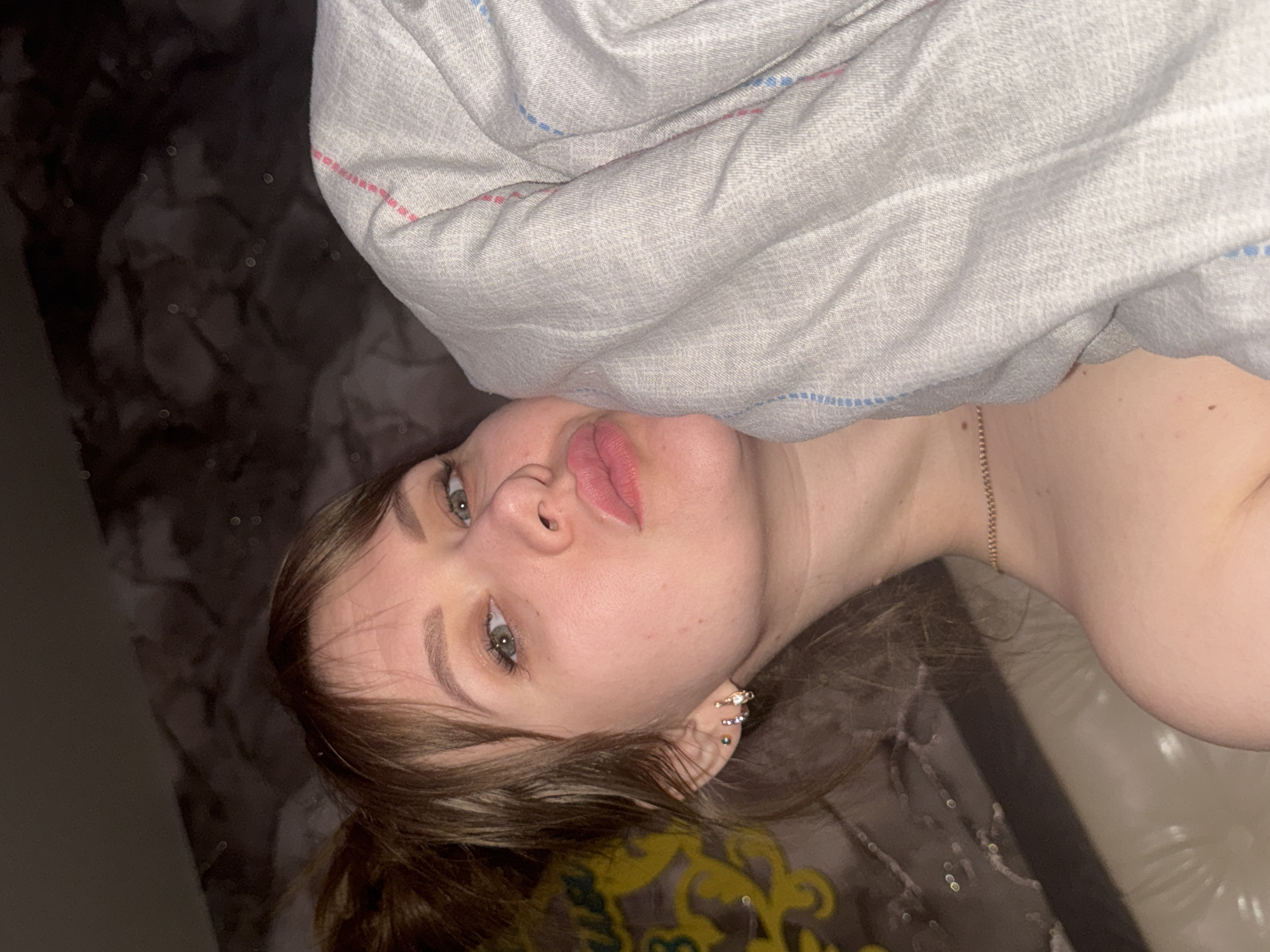The image features a close-up portrait of a young woman, likely in an indoor setting. She is positioned lying down, with her head resting on a surface and her body partially covered by a light grey blanket or duvet with thin, alternating red and blue stripes. The woman has light skin, long brown hair that frames her face, and striking green eyes. Her eyebrows are well-defined, and she wears pink lipstick on her full lips, which are slightly parted. She has multiple ear piercings on her right ear, including a decorative ear cuff with small, possibly jeweled, elements. A delicate gold or rose gold chain necklace is visible around her neck.
She is looking directly at the camera with a composed, neutral expression. The background behind her head is dark and textured, with subtle, reflective patterns suggesting a patterned bedspread, floor, or wall covering. The lighting in the image is dim, indicating an evening or nighttime setting, or a dimly lit room. There are no explicit location cues within the image that point to Chaikovsky, Russia. No visible text is present in the image.