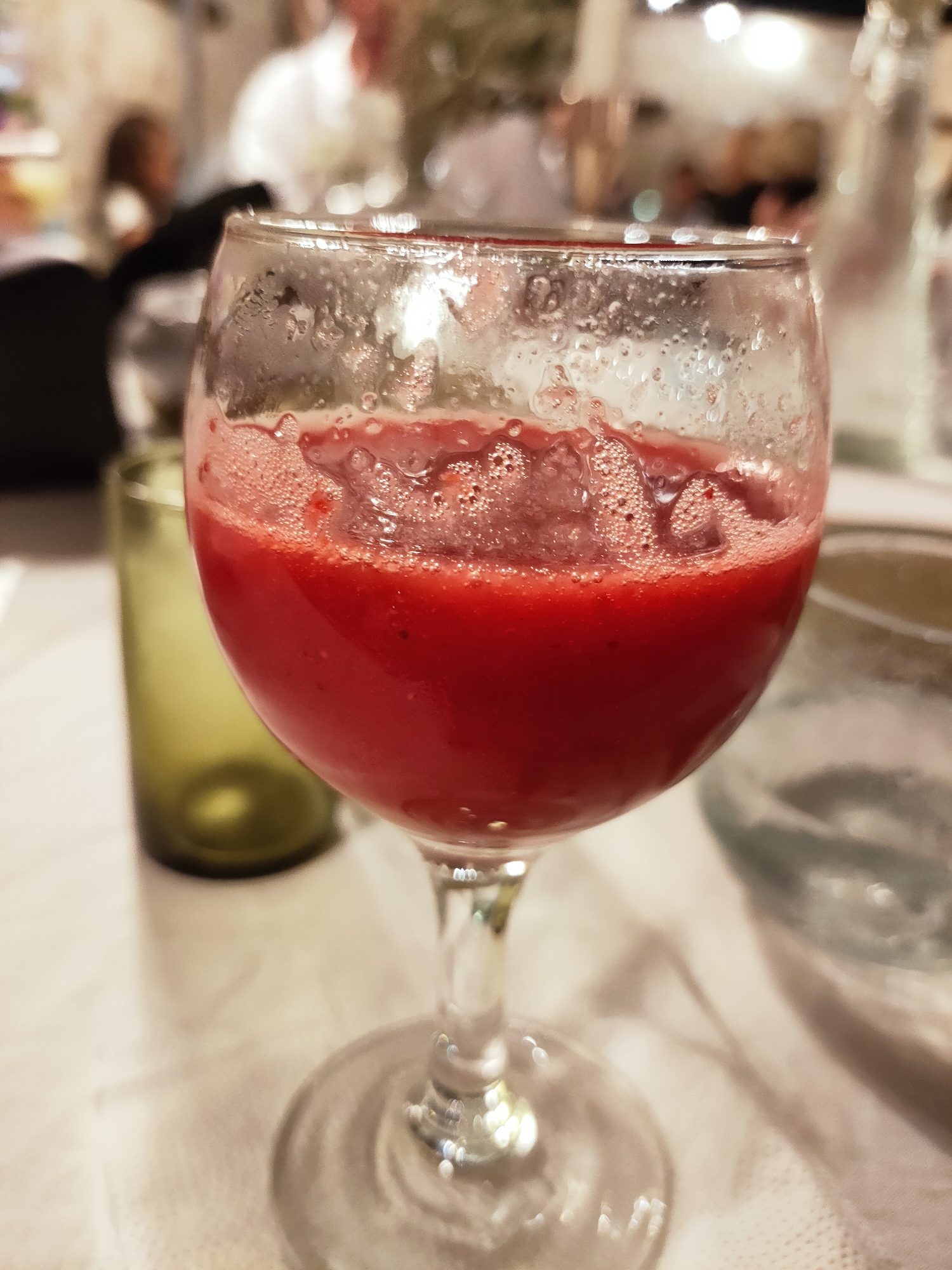 A close-up, indoor, eye-level shot shows a stemmed glass filled with a frothy, red liquid, likely a fruit smoothie or juice, on a white tablecloth. The glass has condensation on its exterior. In the background, slightly out of focus, are other people seated at tables, suggesting a restaurant or event setting in Cebalat, Tunisia. To the left of the main glass, a tall, green glass is visible, also on the tablecloth. The lighting is soft, creating a warm ambiance. The focus is entirely on the red drink.
