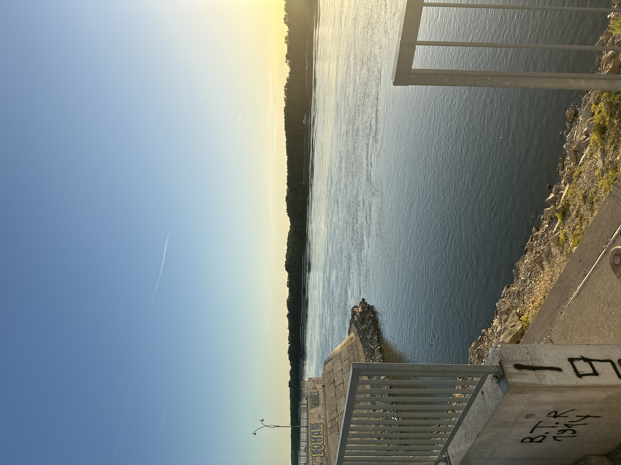 The image captures a serene view of the Danube River at sunset, likely from a vantage point in Ruse, Bulgaria. The sky is a clear, soft blue with a gradient towards a pale yellow on the right, indicating the setting sun. A faint contrail from an airplane is visible across the sky. The river's surface is calm, reflecting the light and showing gentle ripples.
In the foreground, a concrete embankment lines the river. A metal railing runs along the top of the embankment, offering a safety barrier. The concrete structure is adorned with graffiti, including "B.T.R 1914" and some unintelligible markings. Rocky debris and patches of green vegetation are visible along the river's edge, transitioning from the embankment. On the left side of the image, a lamppost with two lights stands on the elevated section of the embankment.
Across the river, a dense line of trees forms the opposite bank. A long, dark structure, possibly a barge or a series of connected boats, is visible on the water near the far bank, silhouetted against the tree line. The overall scene suggests a peaceful evening by the river, with elements of urban infrastructure and natural landscape coexisting.