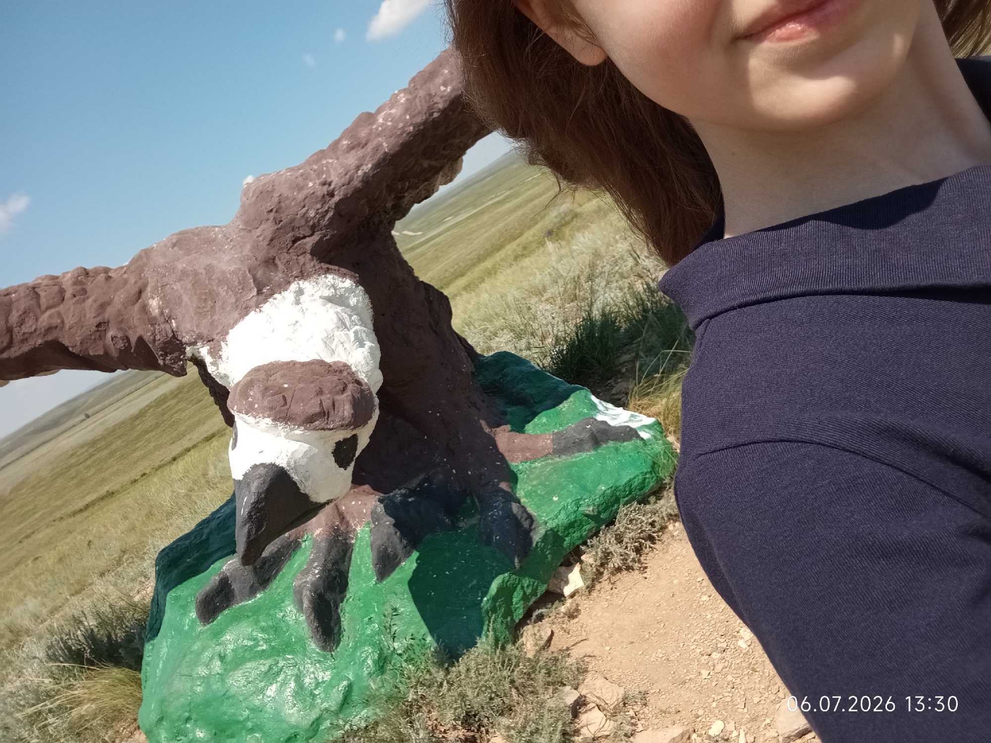 A young girl with long brown hair is taking a selfie outdoors. She is wearing a dark blue collared shirt. Part of her face, including her mouth and chin, is visible in the upper right portion of the frame. In the foreground, there is a brightly painted concrete sculpture of a bird, possibly an eagle, perched on a green, rock-like base. The bird sculpture has brown, white, and black coloring. Behind the sculpture, a vast, grassy plain stretches towards the horizon under a clear blue sky with a few scattered white clouds. The terrain appears dry and arid. The image is captured on a sunny day, suggesting it is daytime. A timestamp "06.07.2026 13:30" is visible in the lower right corner of the image, indicating the date and time the photo was taken. The setting appears to be a rural or remote area, consistent with the landscape of Aleksandrovka, Kazakhstan. The overall mood is casual and relaxed.