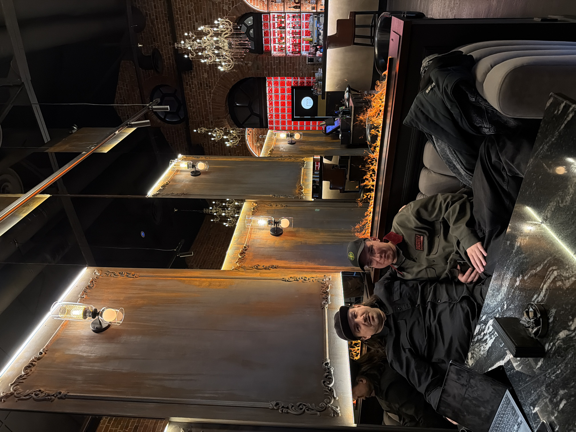 Two men are seated at a table in a dimly lit interior, likely a restaurant or bar. The man closest to the camera is wearing a black shirt and a hat. The second man is wearing an olive green jacket and a hat. A laptop sits open on the table, next to other small items. The table has a dark, polished surface. The walls are decorated with mirrored panels that have ornate frames, with light fixtures placed on the panels. Overhead chandeliers hang from the ceiling. In the background is a bar area with shelves of bottles and decorative red accents. A decorative fireplace with simulated flames is also visible. Based on the architectural style and decor, this image may be from a location in Moscow, Russia.