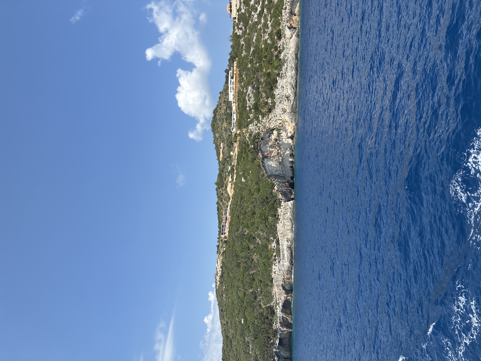 The image depicts a view of a coastal landscape with a body of water in the foreground and a green, hilly terrain with cliffs and scattered buildings in the background. The water is a deep blue with some whitecaps and foam visible on the left side, suggesting movement. The land is covered with dense green vegetation, characteristic of Mediterranean flora, and features rocky cliffs and outcrops. Several buildings, appearing to be modern villas or resorts, are situated on the higher slopes of the hills, some with white facades and red-tiled roofs. The sky is a clear blue with a few scattered white clouds. The scene suggests a sunny day, likely in a coastal resort area. This location is likely within the Elaties Municipal Unit in Greece, known for its scenic coastlines and Mediterranean vegetation. No people or specific actions are clearly identifiable in the image.