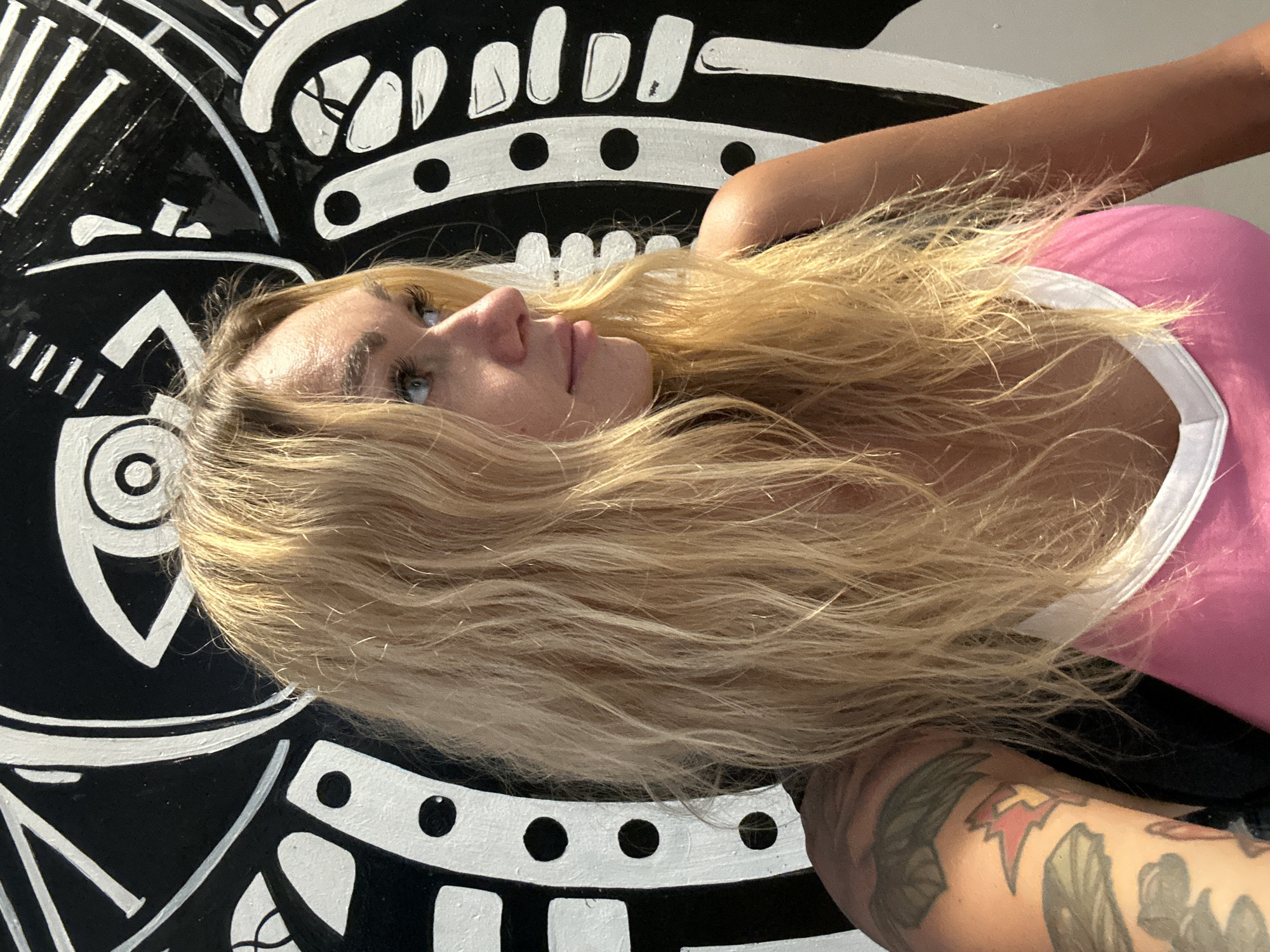 A woman with blonde hair is posing in front of a black and white mural. The mural features abstract designs and an eye-like symbol. She is wearing a pink top with white trim. Her left arm, visible on the left side of the image, is tattooed. She is looking to her right, with her head slightly tilted. The lighting suggests it is daytime. The background mural is an artistic installation. This scene does not appear to be located in Cam Ly - Da Lat, Vietnam.