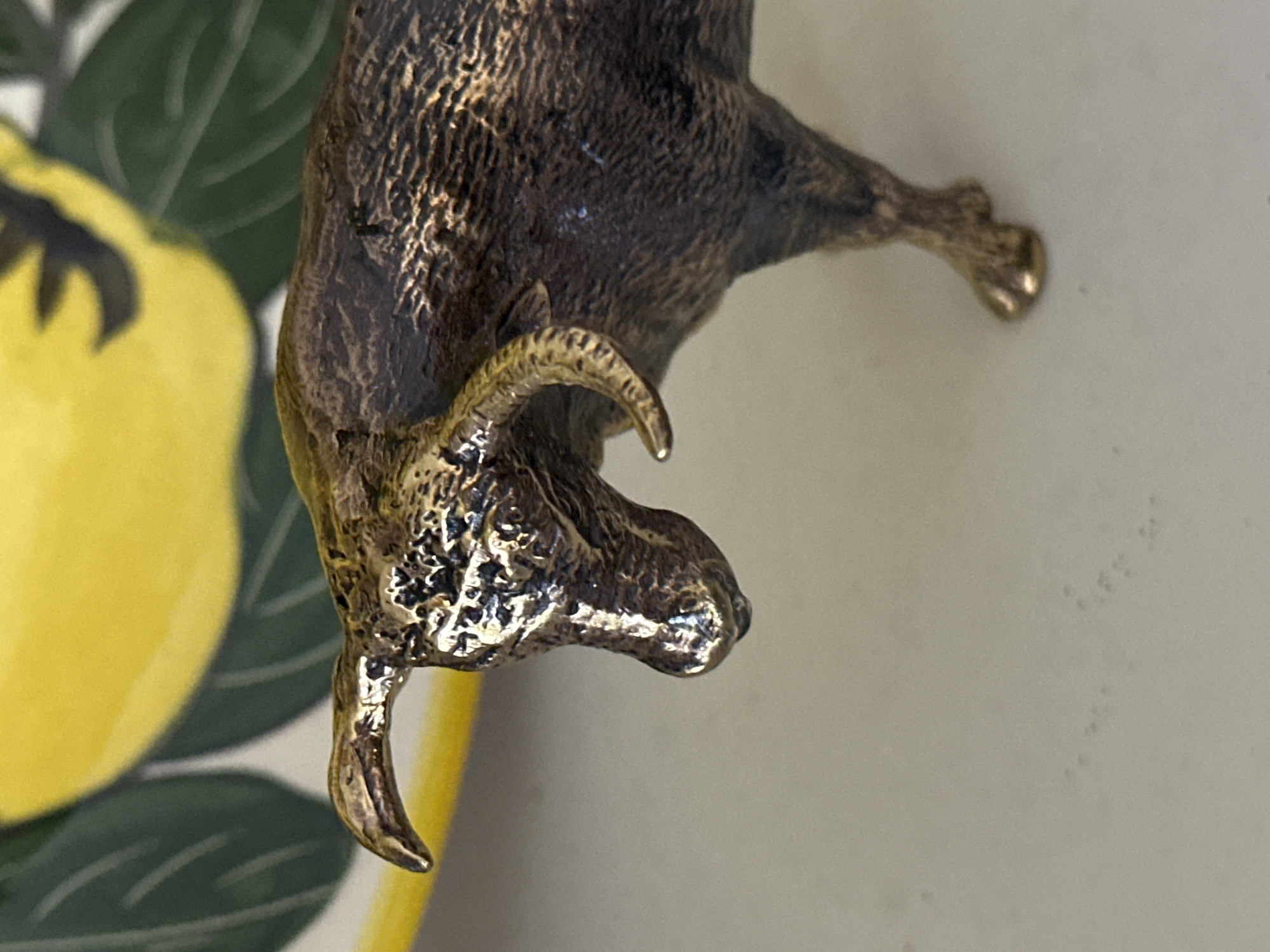 A close-up, eye-level, medium shot captures a detailed view of a small, bronze bull figurine positioned on a light-colored, textured surface. The figurine is oriented to the right and its head is lowered as if grazing. The bull's body displays intricate texturing that mimics the appearance of fur, with a darker patina in the crevices and lighter highlights on the raised areas. Its horns are curved and also exhibit a textured, metallic finish. The bull's legs are visible, with the front right leg appearing to be in focus and the other legs slightly blurred due to the shallow depth of field.
In the background, slightly out of focus, is a portion of a ceramic object. This object features a bright yellow surface with depicted green leaves and possibly a stylized fruit, such as a lemon. The colors are vibrant, contrasting with the metallic tones of the figurine. The lighting in the scene is direct, creating noticeable highlights and shadows that emphasize the three-dimensional texture of the bull figurine.
The image does not contain any information about the city of Nevinnomyssk, Russia, or any people or specific actions.