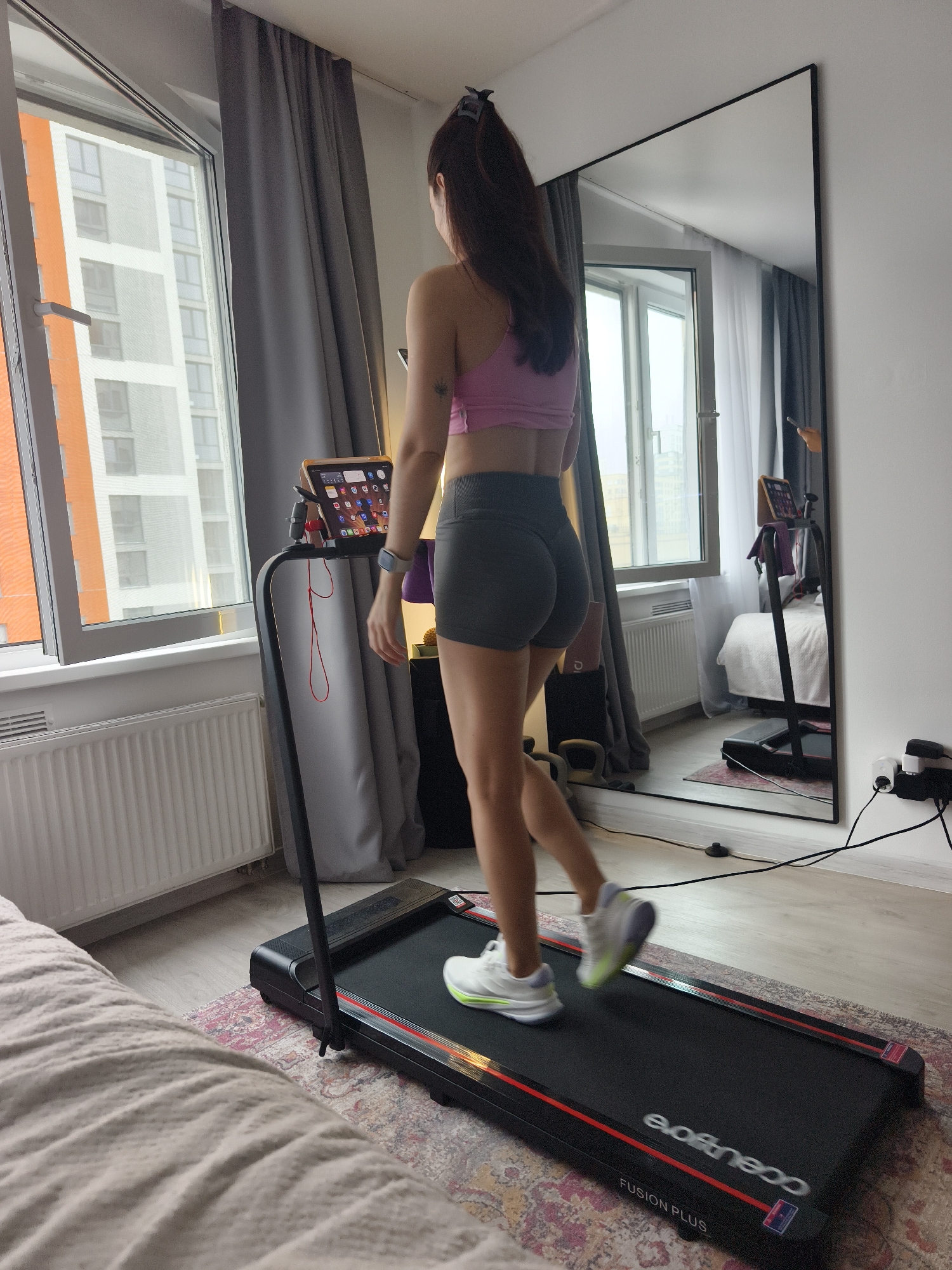 A young woman is shown from behind, exercising on a black treadmill in a well-lit room of a modern apartment in Tyumen, Russia. She has long dark hair tied up and is wearing a pink sports bra or crop top and dark grey athletic shorts. A small tattoo is visible on her upper right arm, and she wears a smartwatch on her left wrist.
The treadmill is black with red accents, bearing the text "FUSION PLUS" and "evoftneco" (which appears reversed, likely "evo fitness"). An electronic tablet, displaying a grid of application icons, is mounted on the treadmill's handle. Red wired headphones hang from the console. The woman's white and green athletic shoes are in motion on the treadmill belt.
Natural daylight enters through a large, partially open window on the left, revealing modern high-rise residential buildings with orange and white facades outside. A white radiator is positioned below the window, and grey curtains are drawn on one side.
A tall, black-framed mirror stands upright on the right, reflecting a segment of the room, including a bed with white bedding, and the treadmill itself. On the wall next to the mirror, a black power strip with white plugs is visible. A patterned rug covers the floor, and a portion of a light-colored bed or sofa is in the bottom left foreground. The scene depicts a home workout setup during daytime in an urban residential setting.
