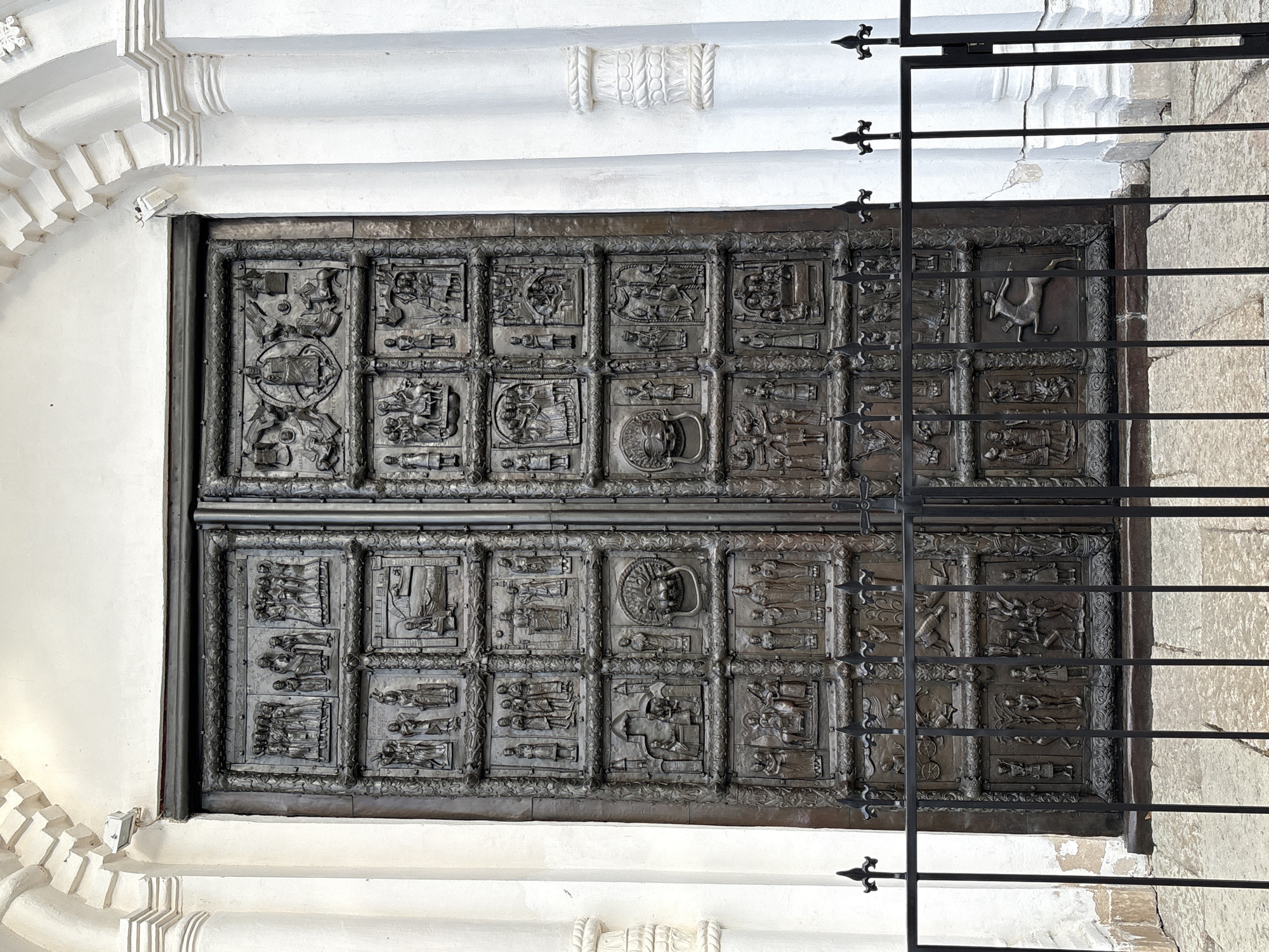 The image showcases the renowned Magdeburg Doors, positioned in the western portal of St. Sophia Cathedral within the city of Veliky Novgorod, Russia. The doors are adorned with intricate bas-relief panels illustrating religious figures and scenes. A metal gate stands in front of the doors, partially obstructing the view. The surrounding architectural details display a white, ornate design.