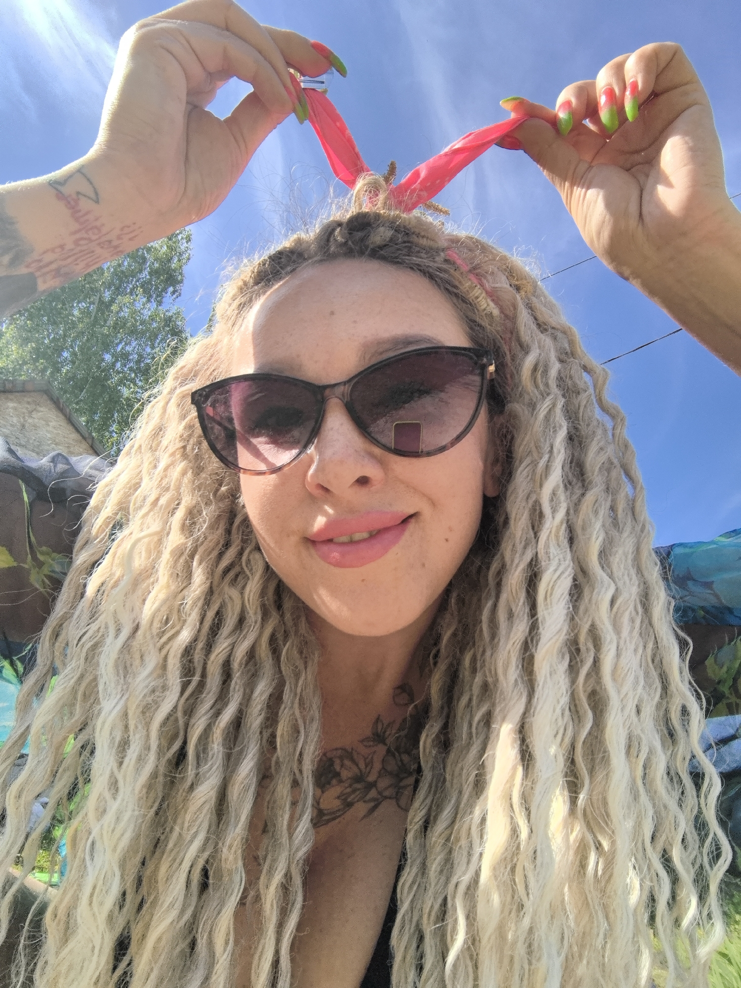 A selfie shot shows a woman with long, blonde, wavy dreadlocks, wearing sunglasses and pink lipstick. Two hands are above her head, holding a pink object that resembles a headband or piece of cloth. The woman's hands have long nails with green tips. Tattoos are visible on the arms and chest. The background features a blue sky, some greenery, and a glimpse of a building. The location is Noviy, Russia.