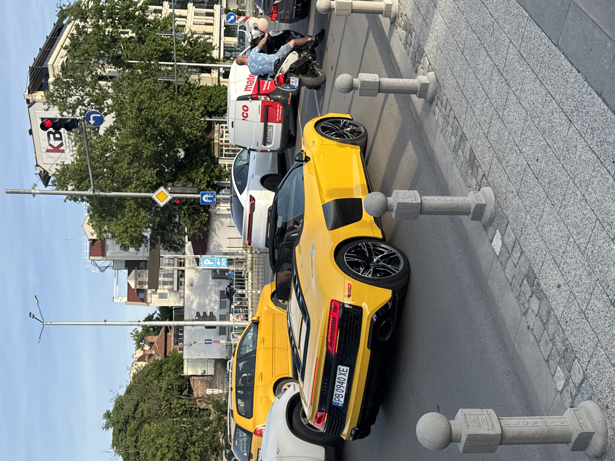 The image shows a street scene in Plovdiv, Bulgaria. A yellow Audi R8 is parked alongside the curb, with its rear facing the viewer. Its license plate reads "PB 0940 XE." Behind the Audi are a white car, a yellow taxi, and a white van with the word "Mateco" on it. A motorcyclist is sitting on their motorcycle. The scene includes street furniture such as bollards made of gray stone and lamp posts. Buildings and trees line the street in the background, and the sky is blue. There are traffic signals and street signs visible.