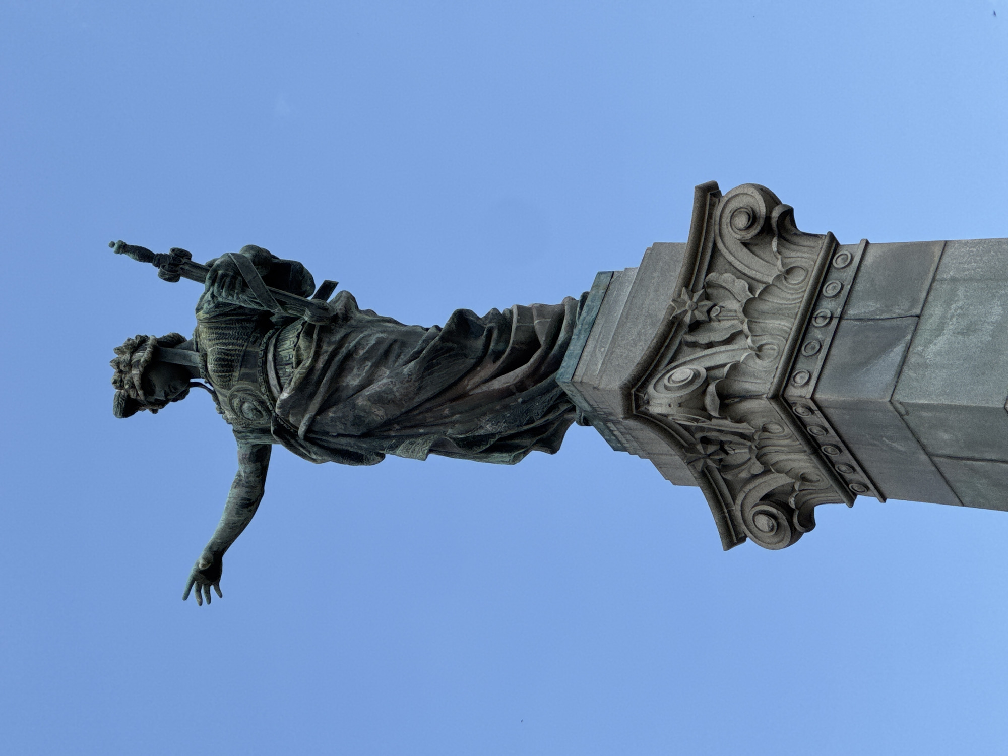 The photograph shows a close-up, low-angle view of a bronze statue of a female figure standing on a stone pedestal. The statue depicts a woman with a crown or headdress, wearing a draped garment and armor. She is holding a sword in her right hand and her left arm is extended to the side with her hand open. The stone pedestal is ornate, featuring carved acanthus leaves and volutes, typical of classical architecture. The statue is positioned against a clear, bright blue sky, with no other elements visible in the background. The lighting suggests it is daytime. This statue is located in the city of Ruse, Bulgaria, and is known as the Monument of Freedom.