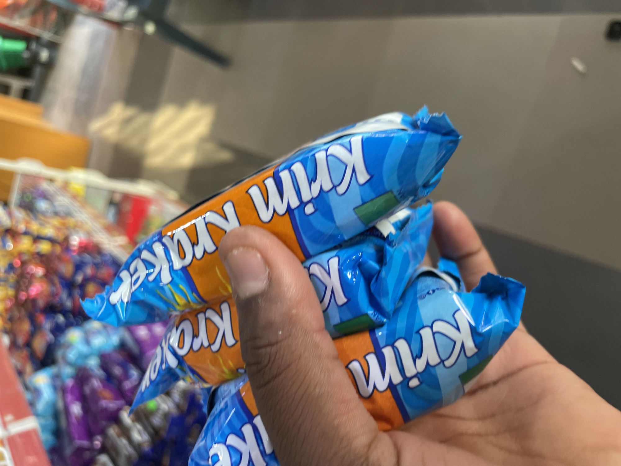 A person is holding three Krim Kraker chocolate bars in their hand. The bars are packaged in blue and orange wrappers. The background shows a store shelf with various items.