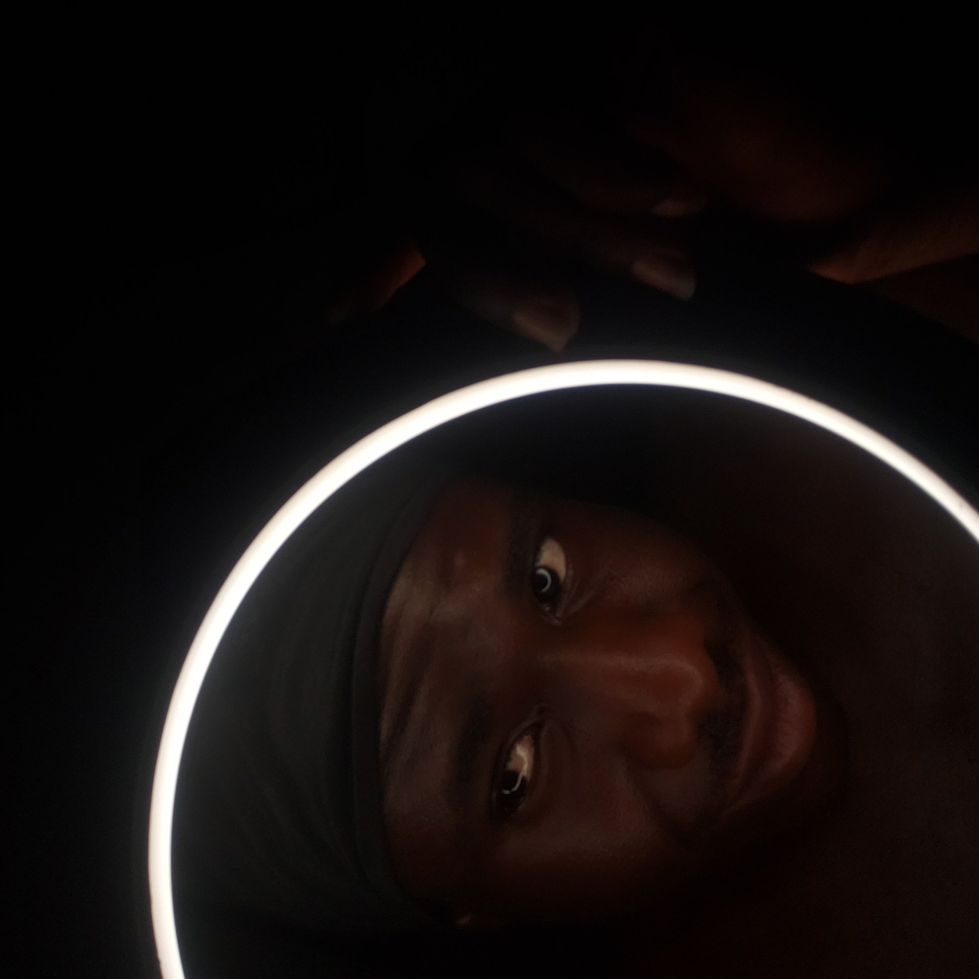A close-up, tilted photograph shows a person's face framed by a ring light against a dark background. The person has dark skin, dark eyes, and is wearing a dark head covering. Their expression appears to be a slight smile. The ring light is positioned around their face, providing the primary source of illumination. The environment is predominantly dark, with the subject's face and the ring light being the focal points. No location within Narayi, Nigeria is evident in the image.