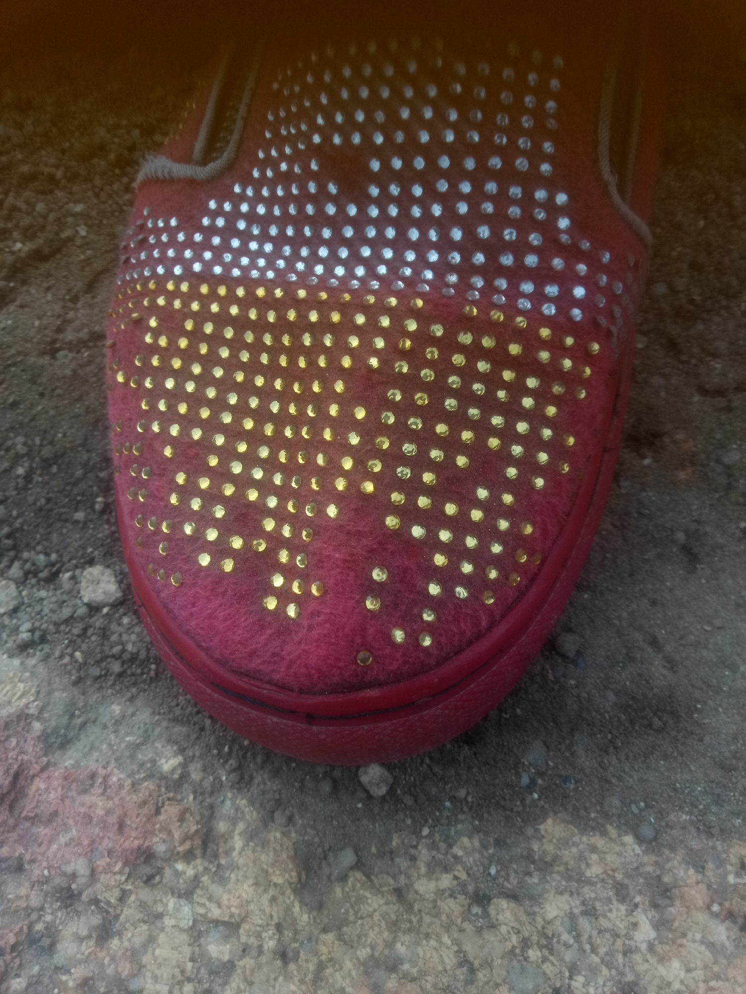 A close-up, overhead shot shows the toe of a bright red slip-on shoe, adorned with a pattern of gold and silver rhinestones. The shoe rests on a textured surface of dirt, gravel, and small rocks, suggesting an outdoor, possibly urban, environment. The lighting casts a warm glow, with some areas of the shoe appearing brighter than others. The focus is sharp on the shoe, with the background slightly blurred, highlighting the detail of the rhinestones and the fabric of the shoe. The scene appears static, with no visible movement or human interaction. The location is not specified but the composition suggests a ground-level perspective.