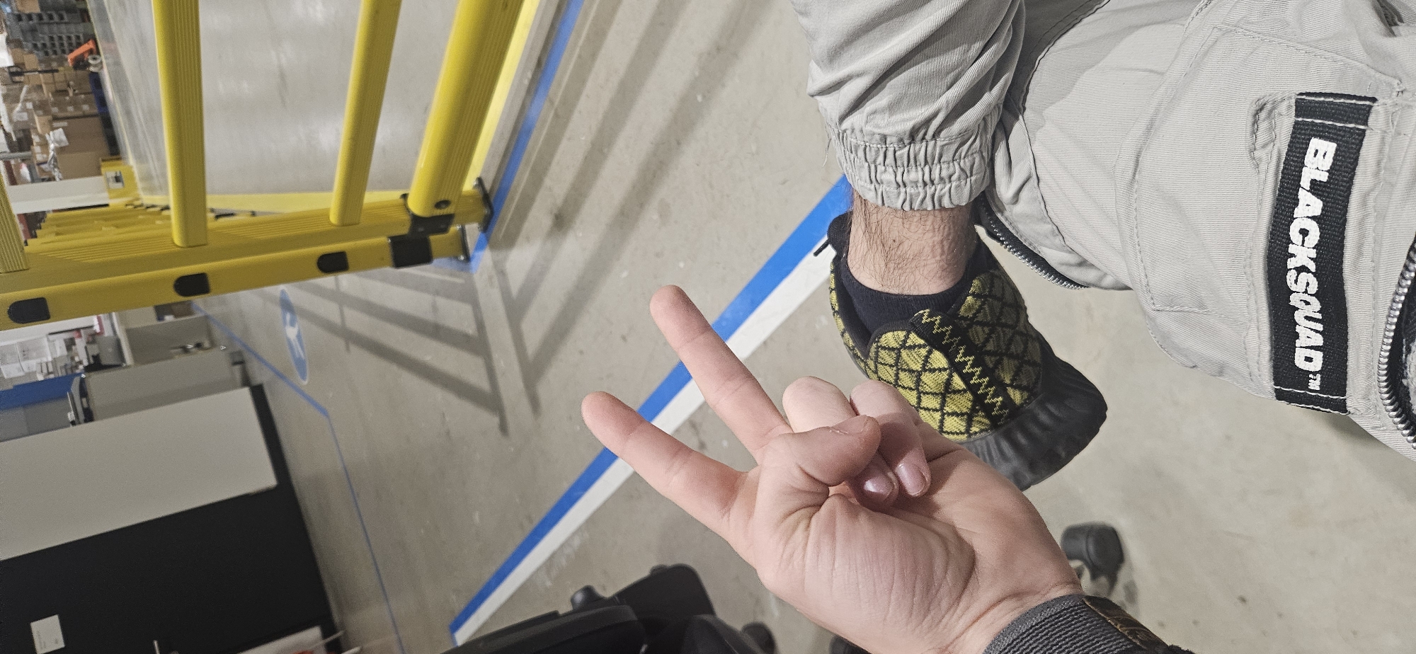 From an elevated perspective, the scene is situated in what appears to be a commercial or industrial interior, potentially a warehouse or retail space. A yellow ladder extends diagonally across the upper left quadrant of the frame. Below, a floor is marked with a blue line.
To the right of the ladder, a person is visible from the knees upward. They are wearing light-colored trousers with gathered cuffs at the ankles, paired with a black and yellow patterned shoe and a black sock. The trousers have a patch on the right leg with the text "BLACKSQUAD".
In the lower center of the image, a hand is extended with the index, middle and pinky fingers raised, while the ring finger and thumb are closed. The person is also wearing a watch. It is unclear what action is being communicated. The identity of the location as being in Plovdiv, Bulgaria is not determinable.