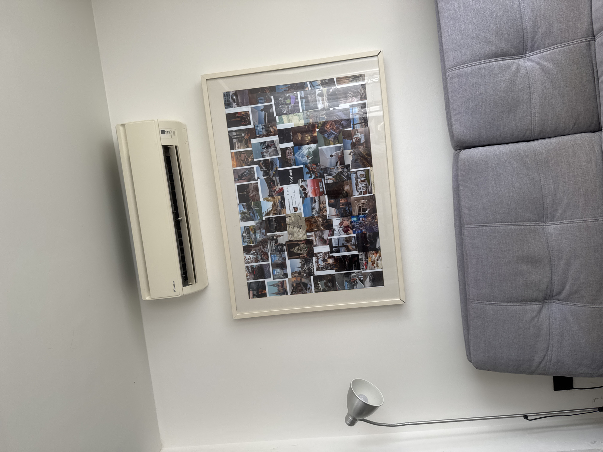The image shows a living room area with a white wall. On the wall, there is a framed collage of many small photographs. To the left of the collage is a cream-colored air conditioning unit. To the right of the collage is a portion of a gray, tufted sofa. Below the collage, on a thin metal arm, is a silver spotlight. The overall impression is that of a well-lit, contemporary interior space. There is no visible text in the image.