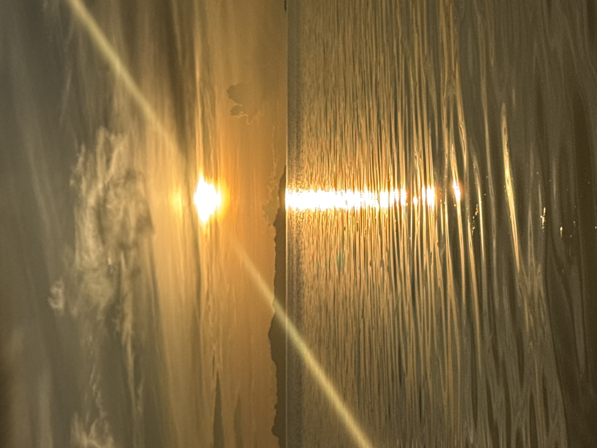 The image depicts a serene ocean sunset with a clear horizon. The sun is positioned in the upper center of the frame, casting a brilliant golden glow across the water and sky. A prominent sunbeam or lens flare streaks diagonally from the top right to the bottom left of the image. The water is calm with gentle ripples reflecting the sunset's colors. In the distance, a silhouetted landmass, likely islands or a coastline, is visible against the horizon. The sky is a blend of warm oranges, yellows, and muted grays, with wispy clouds scattered across it. There are no people or specific objects to note other than the natural elements of the seascape. The location is identified as Baan Pang Ka, Thailand, suggesting this is a scene from that coastal area.