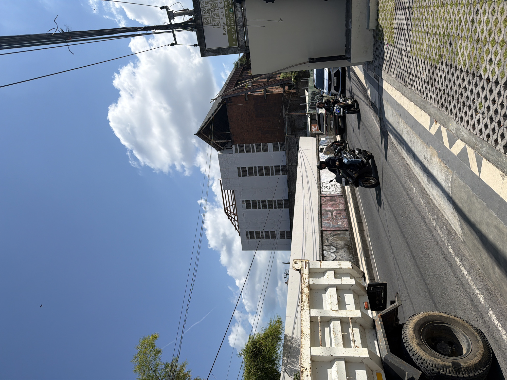 The image, taken in Kuta, Indonesia, depicts a sunlit street scene. The perspective is tilted, making the street appear to ascend diagonally from the bottom right to the middle left.
In the foreground, the rear of a large white dump truck is prominent, with its large tire visible on the right. The street, a paved asphalt road, shows light vehicle traffic. Two individuals on black scooters, both wearing helmets, are visible, suggesting active commuting. Several parked cars are also present along the side of the road.
To the right of the road, a sidewalk features distinct interlocking concrete pavers with grass growing between them. On the left, buildings line the street. One building has a brick upper story with a distinct roofline and a white lower section. Adjacent to it is another white building with multiple small, rectangular windows.
Overhead, a bright blue sky dominates, adorned with large, fluffy white clouds. Numerous power lines and utility poles are tangled against the sky on the left. Green foliage from trees is visible in various parts of the frame, hinting at tropical vegetation.
A white signboard is visible, displaying "SPA" prominently. Below it, smaller text reads "Body Face Na," followed by an address: "Jl. Drupadi No. 1010 S." Phone numbers "P: +62 811 39:33 700 +62" are also legible. A partial license plate "K3769 ACT" and "DK 8140" on a car are also visible. The clear sky and strong shadows indicate it is daytime, likely midday or early afternoon.