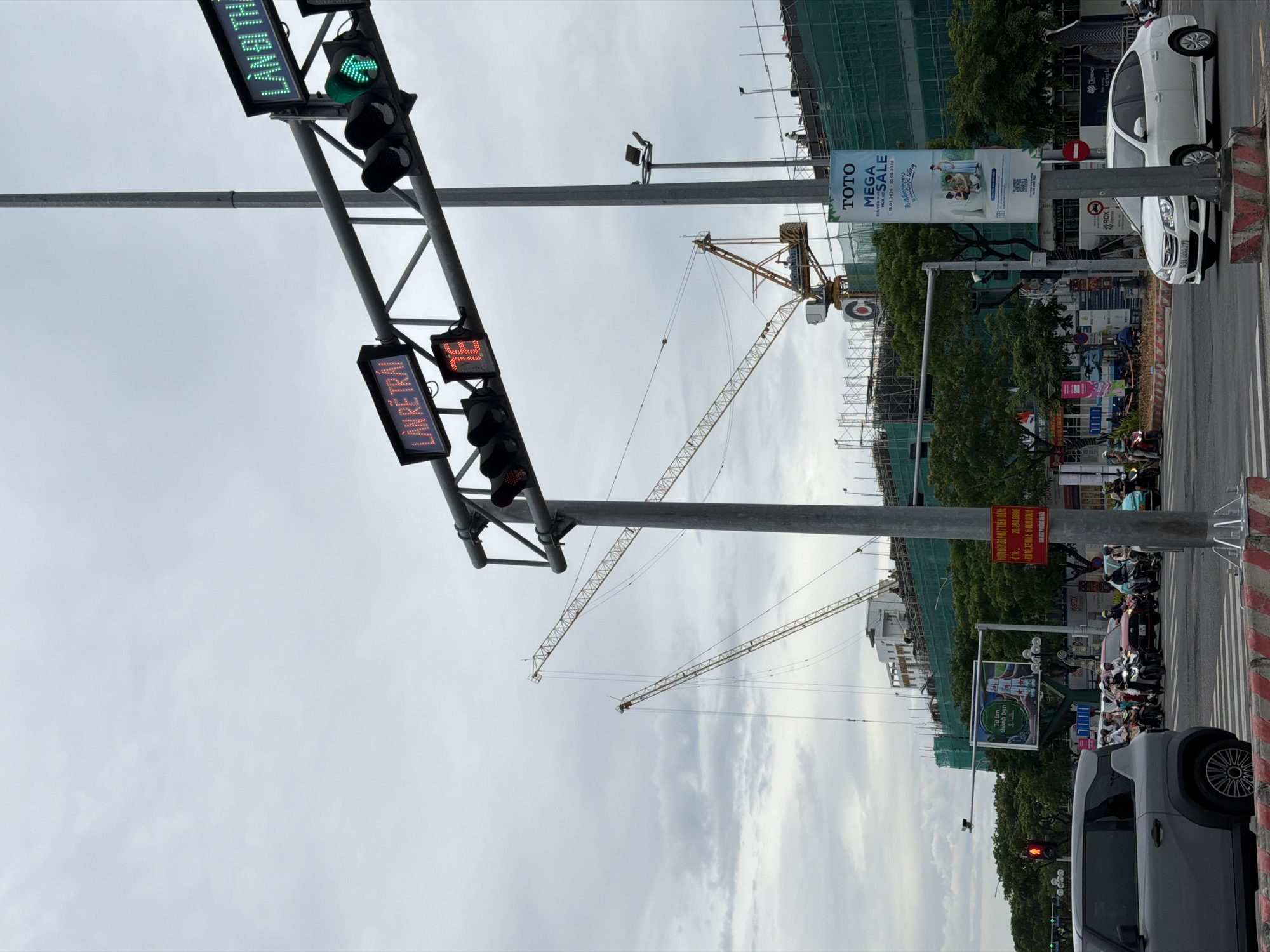 The image captures a street scene in Da Nang, Vietnam, with a focus on traffic infrastructure and urban development. Overhead traffic lights display "LAN RE TRAI" in red and a green arrow pointing left, indicating a lane for left turns. Another digital sign reads "LAN BI THA" in green. A traffic light at the bottom left shows a red signal for vehicles.
Construction is prominent in the background, with several tall cranes towering over a building covered in green netting. Scaffolding is also visible.
In the foreground, traffic moves through an intersection. A grey SUV is parked on the left side of the frame, and a white sedan is turning right. Numerous motorcycles, many with riders wearing helmets and masks, are also present, suggesting typical urban traffic flow.
Several signs are visible. One is a large banner for "TOTO MEGA SALE." Another is a red sign with Vietnamese text. A billboard-like display featuring what appears to be a beer advertisement is also present.
The sky is overcast, with scattered clouds, contributing to a diffused light. Trees line the sides of the street, and various street poles and traffic signals are characteristic of a busy city intersection. The overall impression is one of an active construction site within a bustling urban environment.