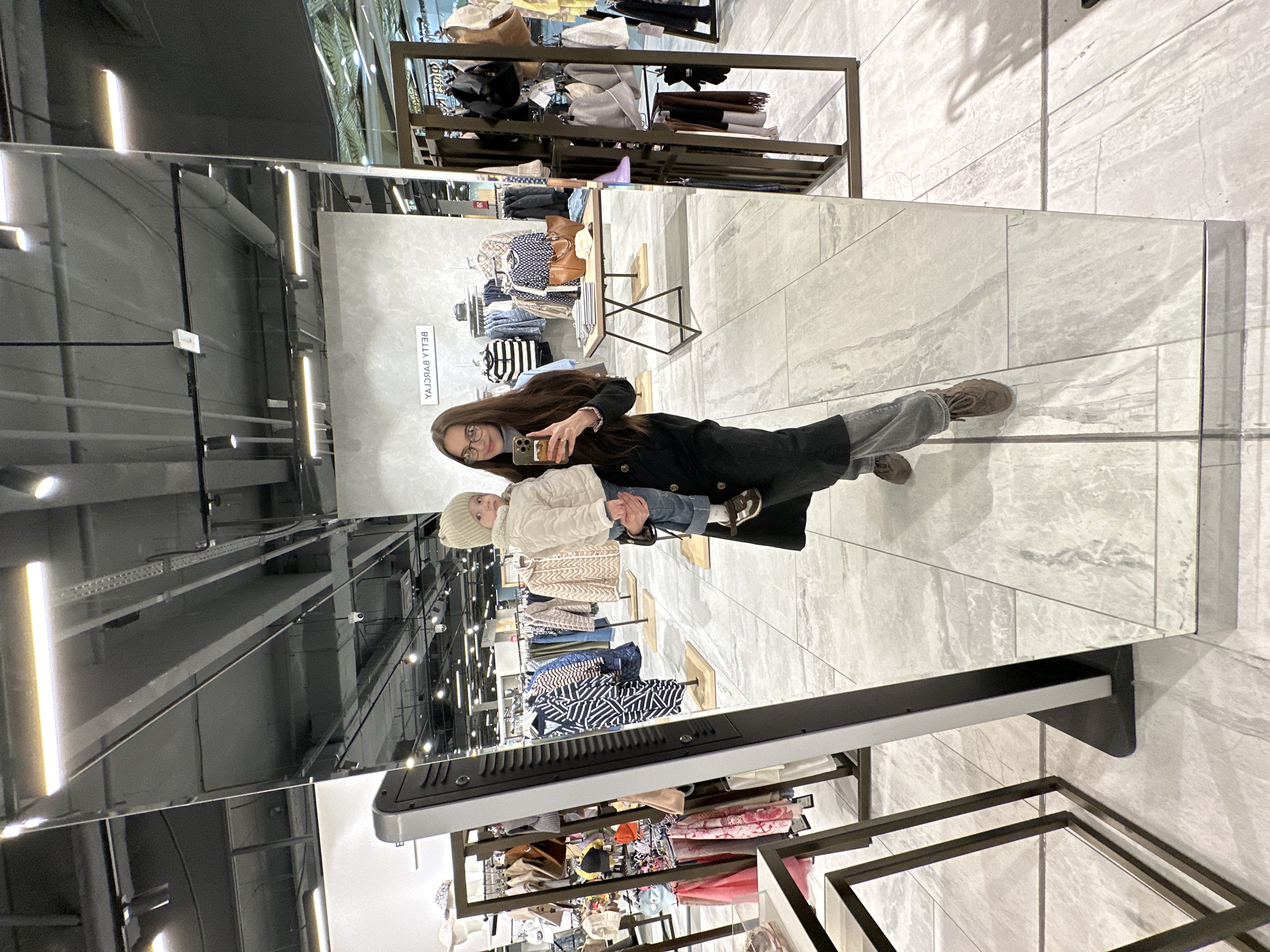 The image depicts a woman holding a young child, both reflected in a large, full-length mirror within what appears to be a modern retail clothing store in Yekaterinburg, Russia. The woman, with long dark hair and wearing glasses, is holding a smartphone in her right hand, seemingly taking a selfie of herself and the child in the mirror. She is dressed in a dark coat, light-colored pants, and brown shoes. The child, held upright by the woman, is wearing a light cream-colored quilted jacket, a matching beanie, blue jeans, and small brown shoes, looking directly at the reflection.
The scene features a clean, tiled floor with light grey rectangular tiles. In the background, both directly and reflected in the mirror, are various clothing racks and display tables laden with folded and hanging apparel, including sweaters, striped shirts, and accessories. The ceiling shows industrial-style lighting fixtures and visible ductwork, indicative of a modern commercial space. A security camera is also visible on the ceiling. Mirrored text, "BELLA BYBCYA", is displayed on a sign or poster visible in the reflection behind the subjects. The overall environment suggests a well-lit, organized retail boutique.