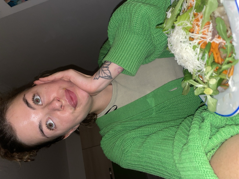 A young adult female, with light eyes and dark hair tied in a bun, is depicted in a dimly lit indoor setting in Boreti, Montenegro. She is wearing a bright green knitted cardigan over a light beige ribbed top. Her left hand is positioned to support her chin, revealing a stylized leaf or petal tattoo on her forearm. She looks upwards and to the right with a contemplative expression. In the immediate foreground, a white plate with a blue rim contains a salad comprised of green leafy vegetables, shredded orange carrots, and a generous topping of grated white cheese. A light-colored wooden cabinet is partially visible in the blurred background to the left.
