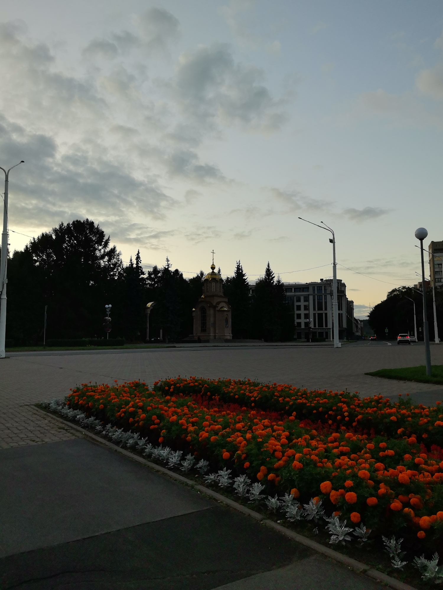 The image depicts a wide, paved plaza in Kemerovo, Russia, at dusk. In the foreground, a raised flower bed is filled with vibrant orange marigolds and bordered by silvery-grey foliage. To the left, a tall lamppost stands near the edge of the plaza.
In the middle ground, a small chapel with a golden dome is centrally located. Behind it, a dense line of dark green trees forms a backdrop. Further back, a modern, multi-story building with a light-colored facade is visible, and beyond that, more trees and buildings line a street where a few cars are driving. Another lamppost is present on the right side of the plaza. The sky above is filled with a mix of grey and light blue clouds, indicating the twilight hour. The overall impression is of a serene urban landscape in the early evening.