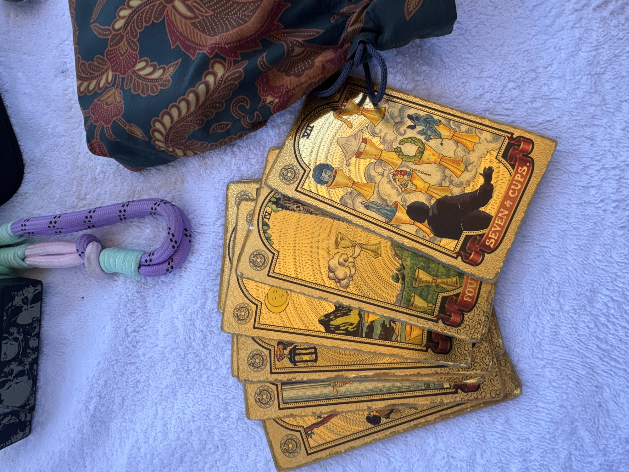 A deck of gold-edged tarot cards rests on a white, textured surface alongside a patterned pouch, a looped rope, and a black object. The topmost tarot card displays the text "SEVEN 4 CUPS." The other cards are partially obscured, but reveal additional suits and numerals like "IV." The pouch is dark green with a brown and red paisley-like pattern and is tied with a blue drawstring. The rope is purple, white, and mint green, knotted in a loop. The black object is only partially visible on the left edge of the image. The background is the white, textured surface.