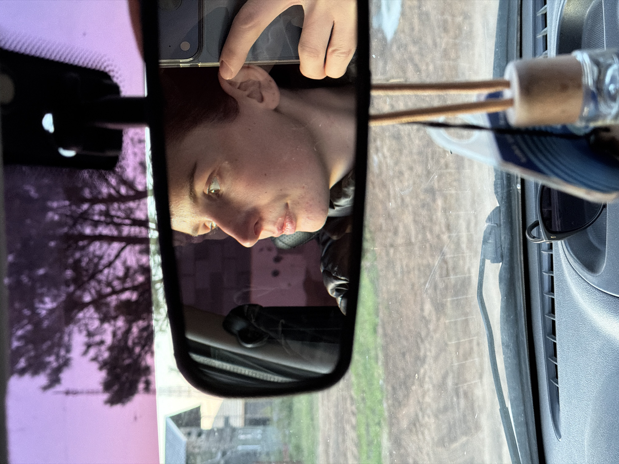 The image is taken from inside a vehicle, looking through the windshield and rear-view mirror. The rear-view mirror reflects a person holding up a phone, possibly taking the picture. The person has fair skin and short, reddish-brown hair. Their face is partially visible in profile. Outside the car, a landscape is visible through both the rear-view mirror and windshield. Details are indistinct, but it appears to be a residential area with buildings and trees. The lighting indicates it is daytime. An air freshener with wooden sticks hangs from the rear-view mirror. The interior of the car is visible around the mirror. There is no definitive evidence to confirm that the location is in Chaikovsky, Russia.