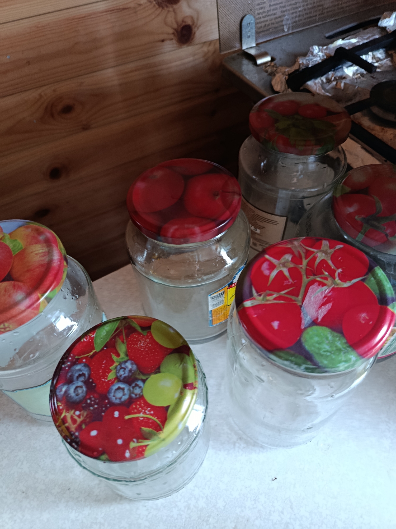 The image shows a collection of six empty glass jars with decorative lids, arranged on a white surface, likely a countertop or table. The jars are of varying sizes and are positioned in a cluster, with some partially overlapping. The lids are adorned with colorful patterns of fruits and vegetables. One lid features red apples, another has red tomatoes and green leaves, and a third displays strawberries, blueberries, and grapes. The background consists of a wooden paneled wall, suggesting an indoor setting. To the right, a portion of a kitchen stove with metallic surfaces and black burners is visible, indicating the jars are likely in a kitchen environment, possibly in the city of Верея, Russia. There are no people or animals present in the image. The overall scene is static and depicts stored items.