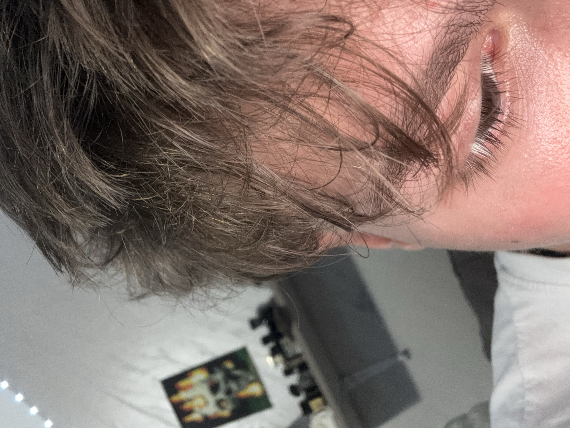A close-up, tilted view of a person's face and hair. The person has light brown hair and fair skin. One eye is visible, with dark eyelashes. The person is wearing a white shirt. In the blurred background, there is a wall with a poster and some equipment. The location in the city of Troyan, Bulgaria, is unknown.