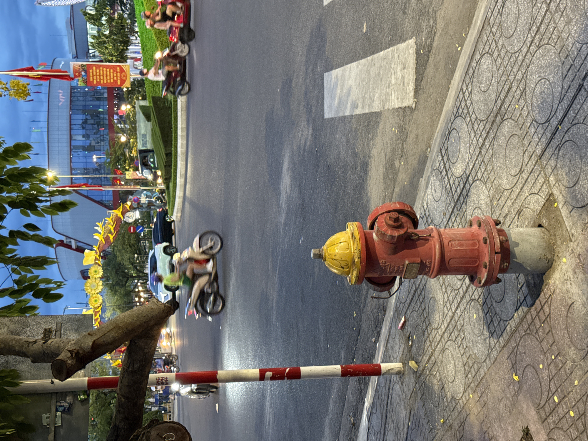 This image captures a street scene in Nha Trang, Vietnam, likely during the early evening or late afternoon given the lighting. In the foreground, a red fire hydrant with a yellow top stands on a patterned concrete sidewalk. Adjacent to the hydrant is a red and white striped pole. The sidewalk is littered with a few small yellow flower petals.
Beyond the sidewalk, a paved road shows signs of recent rain with reflective patches. A white pedestrian crossing is visible on the road. Several motorcycles are on the road; one cream-colored motorcycle with a rider wearing a green shirt and shorts is in motion. Further in the background, two people are riding a red motorcycle.
The middle ground features a traffic circle with neatly trimmed green bushes and a small monument. A modern building with a reflective glass facade dominates the background. Red and yellow flags, some bearing Vietnamese text, are displayed on poles in front of the building. Decorative yellow floral garlands are suspended overhead, illuminated by streetlights. The sky is a dusky blue, indicating twilight. The overall scene depicts a well-maintained urban environment with active traffic.