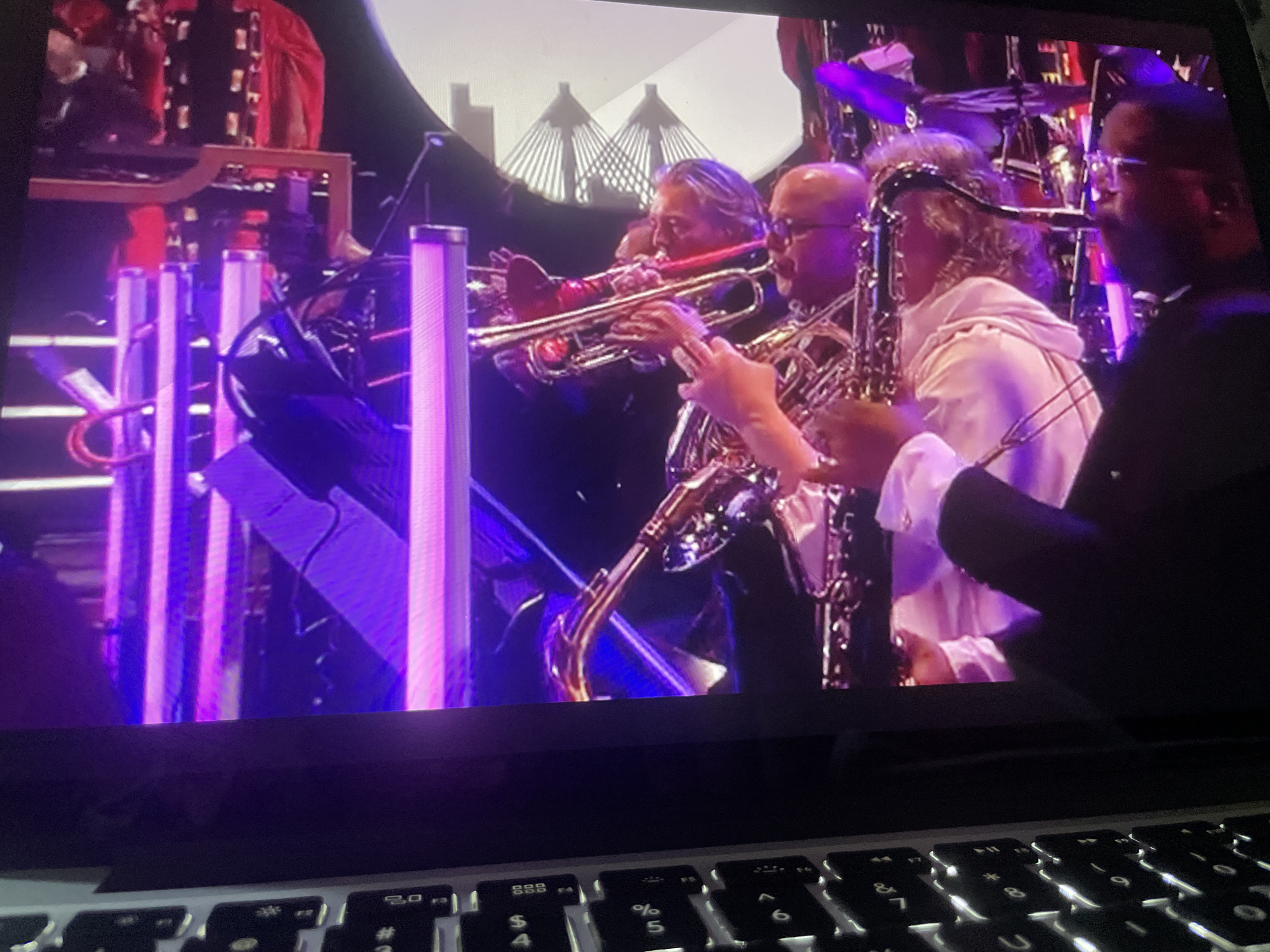 The image shows a screen displaying a live music performance. In the foreground, a laptop keyboard is visible. The performance features a band with several musicians playing brass instruments. Two men are prominently featured, one playing a trumpet and another playing a saxophone. Another musician, partially obscured, is also seen playing a saxophone. The stage is lit with vibrant purple and pink lights, and there are vertical light fixtures. In the background, a blurred image of a cityscape with distinctive bridges can be seen. The overall impression is that of a concert or event taking place in a venue with stage lighting. The location in Howayi, Nigeria is not evident from the visual information provided.
