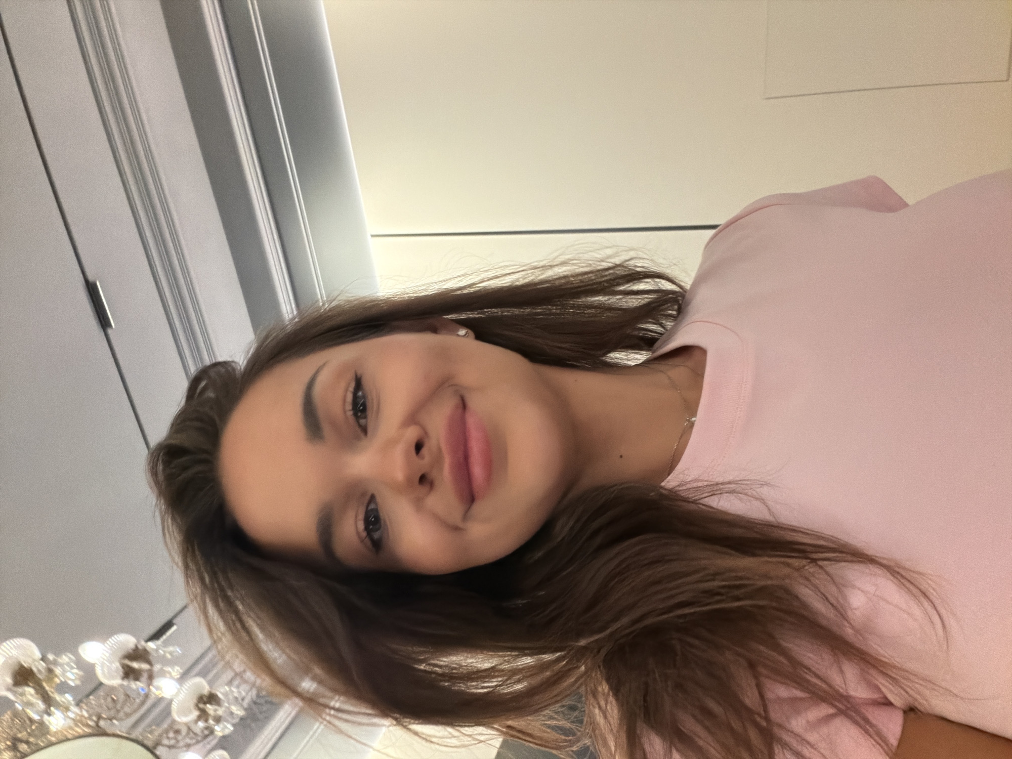 A young woman with long, brown hair and blue eyes is captured in a close-up, selfie-style shot. She is wearing a light pink t-shirt and a delicate necklace. Her hair is slightly windswept, framing her face. She has a warm smile, and a small stud earring is visible on her left ear. The background features a portion of a white ceiling with ornate light fixtures, possibly a chandelier, and a contemporary-style ceiling with recessed lighting. The right side of the image shows a plain, off-white wall, possibly a closet door or a wall panel. The lighting appears to be artificial. The scene is indoors, and the setting suggests a residential or hotel interior. There are no other people visible in the image.