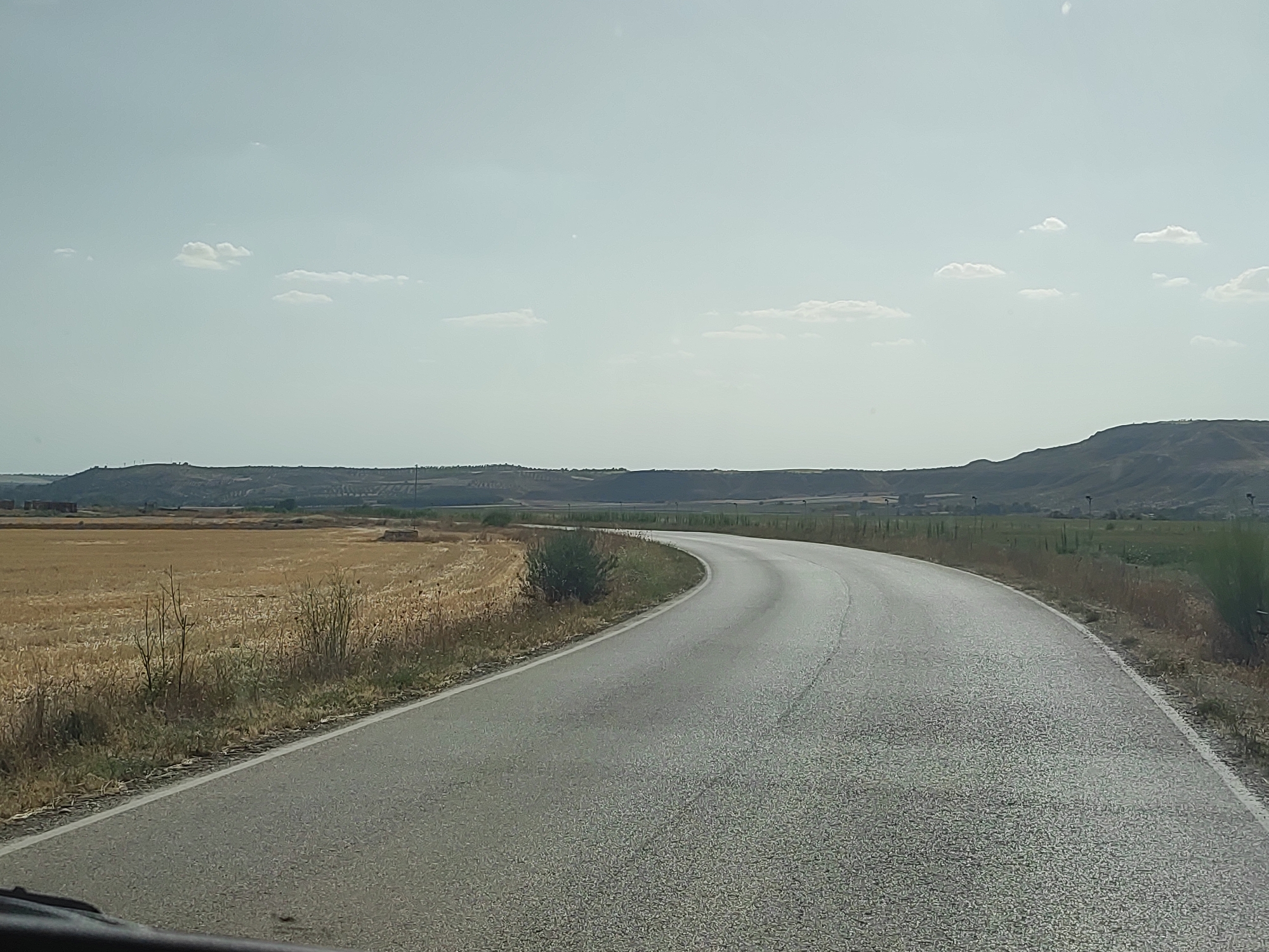 The image depicts a rural landscape in Illana, Spain, dominated by a winding asphalt road curving from the lower left to the mid-right of the frame. The road surface appears worn, with visible texture and occasional darker patches. To the left of the road, a field of dry, golden stubble or hay stretches towards the horizon, bordered by sparse, dry vegetation and a few small, leafless trees. In the distance, a gentle, undulating landscape of hills forms the horizon, partially obscured by a hazy, pale blue sky with scattered, wispy white clouds. The overall impression is one of a dry, sunny day in a rural, agricultural area.