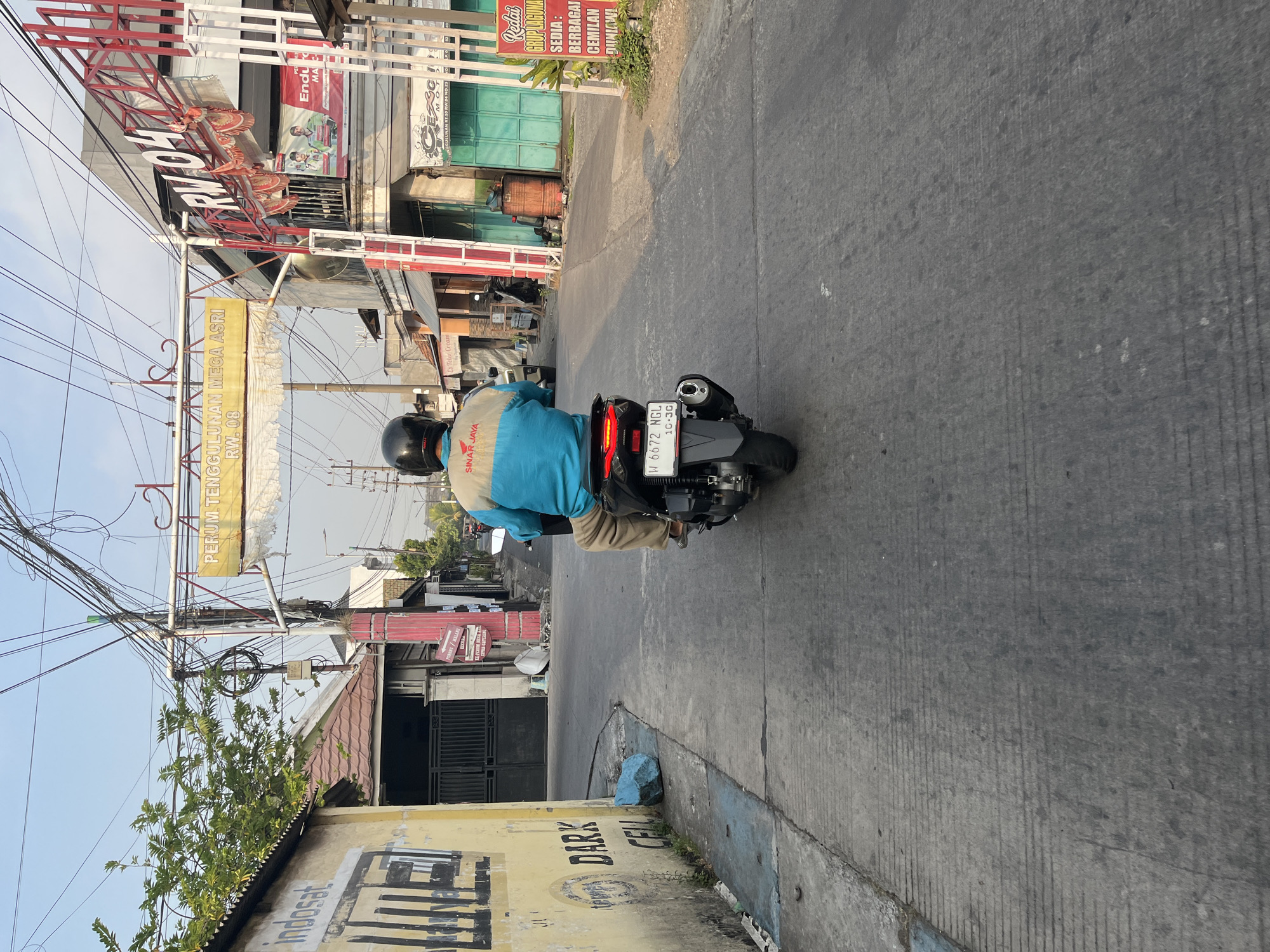 The image shows a person riding a motorcycle down a street in Tenggulunan, Indonesia. The motorcycle has a license plate that reads "W 6672 NGL 10-30". The rider is wearing a black helmet and a blue and tan jacket with "SINAR JAYA" written on the back. The street is paved and has some cracks and lines. On the left side of the street, there is a yellow building with "indosat" and "DARK CELL" written on it. On the right side of the street, there are several buildings and shops, some of which have signs in Indonesian. There is an archway overhead with "PERUM TENGGULUNAN MEGA ASRI RW.08" written on it, and a sign that says "RW 04". The sky is clear and blue, and the sun appears to be low in the sky, casting shadows. There are several utility poles and wires overhead.