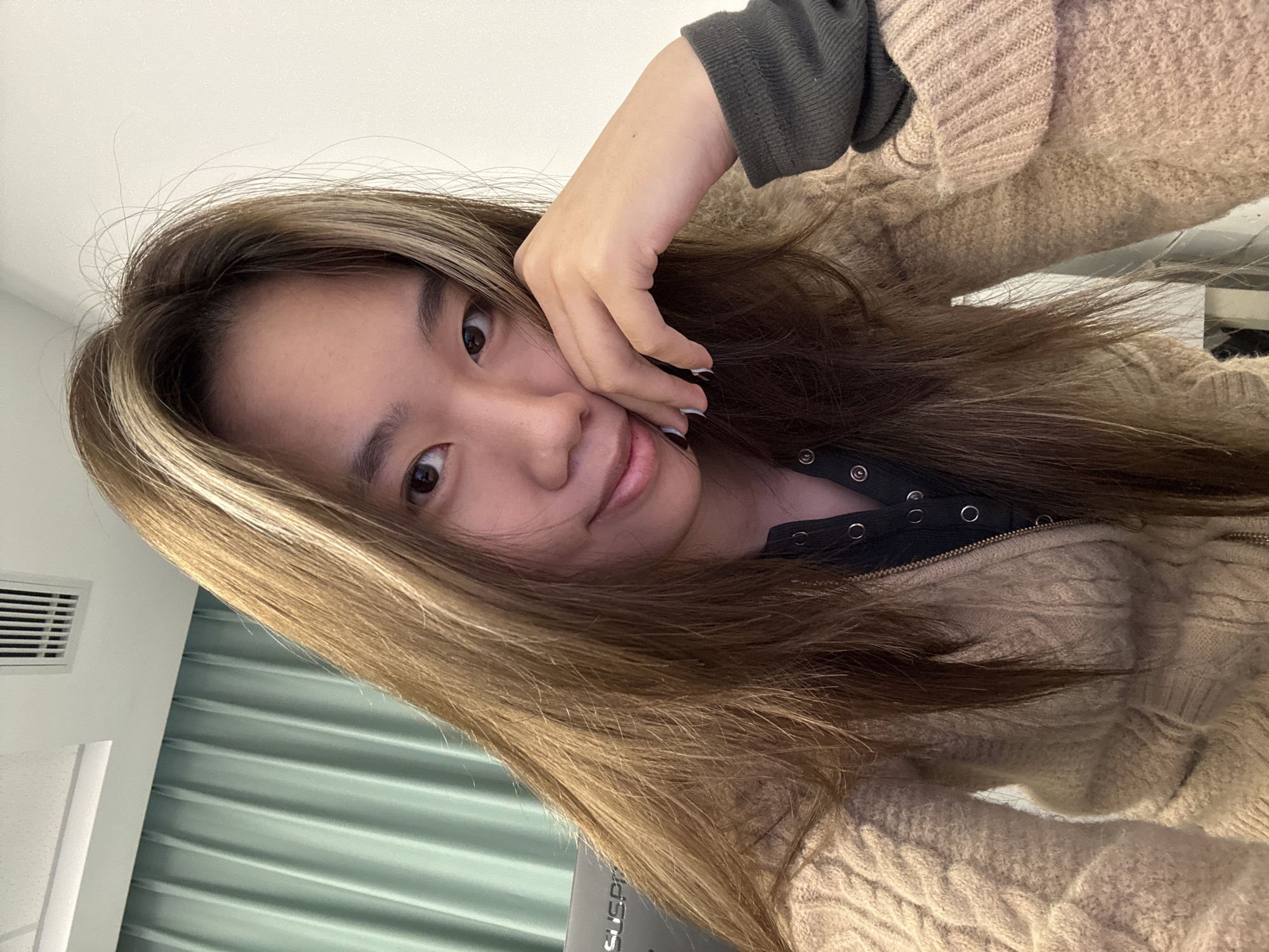 A young individual with light brown hair with blonde highlights is looking at the camera and touching their face with their right hand. They are wearing a dark grey shirt underneath a light brown, cable-knit sweater. Background shows a wall with pale green, corrugated panels and an air vent in the upper left corner. The location is unspecified within Nan'an District, China, as the image does not provide identifying locational features.