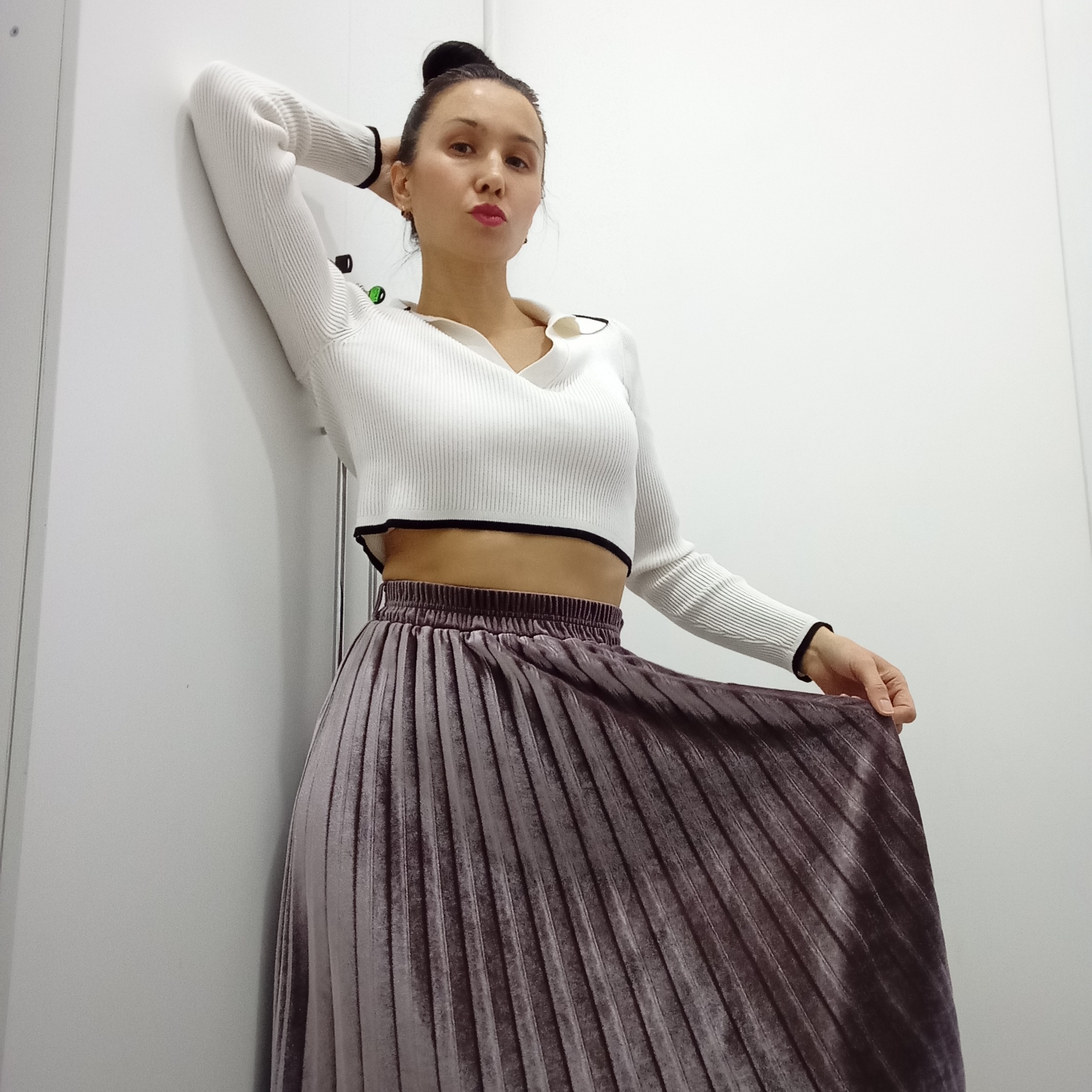A woman is captured in a medium shot, leaning against a plain white wall in what appears to be an indoor setting. She stands with her body slightly angled, her left arm raised and bent, resting her hand behind her head. Her right hand gently holds a portion of her pleated skirt.
She is wearing a white, ribbed knit crop top with long sleeves, featuring a collar and black trim along the cuffs and hemline. Her midriff is visible between the crop top and the high-waisted, pleated velvet skirt, which is a muted mauve or dark grey color. Her dark hair is styled in a bun on top of her head, and she has small earrings. She looks directly at the camera with a confident expression, her lips slightly pursed.
The background consists of a stark white wall, with a faint vertical line on the left side suggesting a door frame or panel. The lighting is even and bright, highlighting the textures of her clothing and the simplicity of the setting. There is no visible text in the image. This scene takes place in Izhevsk, Russia.