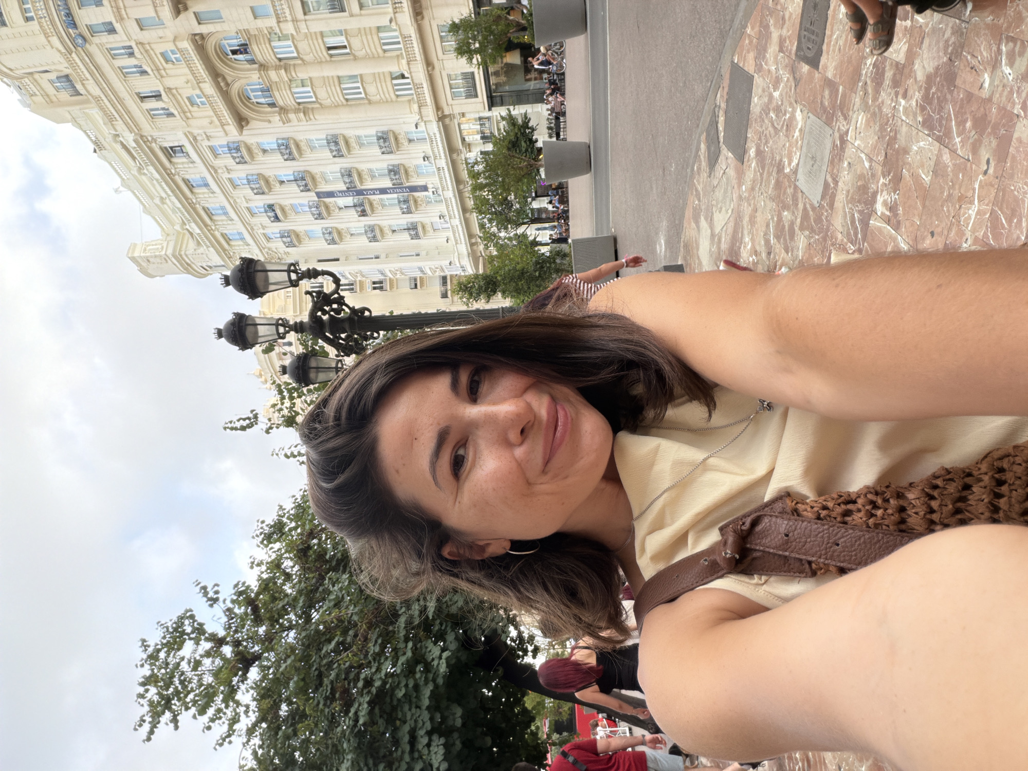 A woman is taking a selfie in an outdoor urban setting. She is in the foreground, smiling at the camera. She has dark brown hair, is wearing a light yellow top, and a brown bag is slung over her shoulder. In the background, a large, ornate building with many windows dominates the scene. There are also trees, lampposts, and people walking on a patterned pavement. The environment suggests a city square or a well-trafficked public area.