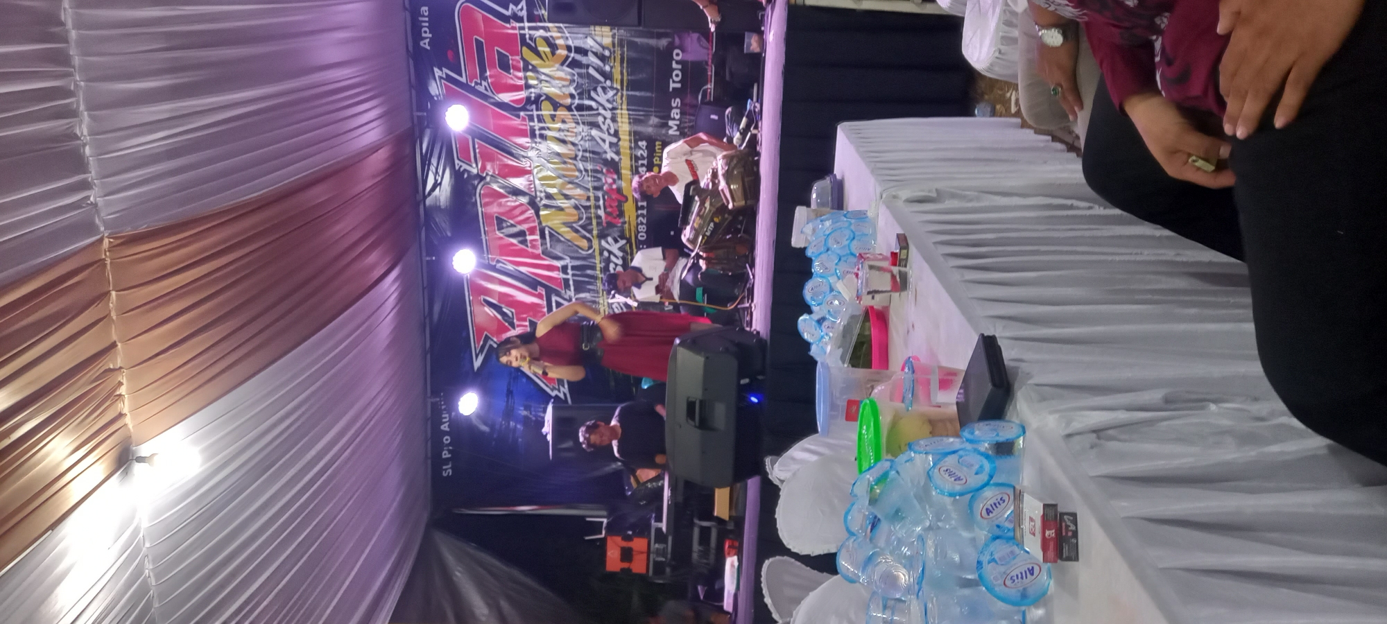 A live musical performance is taking place under a tent in Sidamulya, Indonesia. A female singer in a red dress stands center stage, holding a microphone. She is backed by a band, including a keyboard player to her left and a drummer to her right, all seated. The stage backdrop features a banner with the text "Apila Musik" and phone numbers. In the foreground, a table is laden with plastic cups, water bottles, and cigarette packs, suggesting a casual event. The overall atmosphere appears to be a local gathering or celebration.