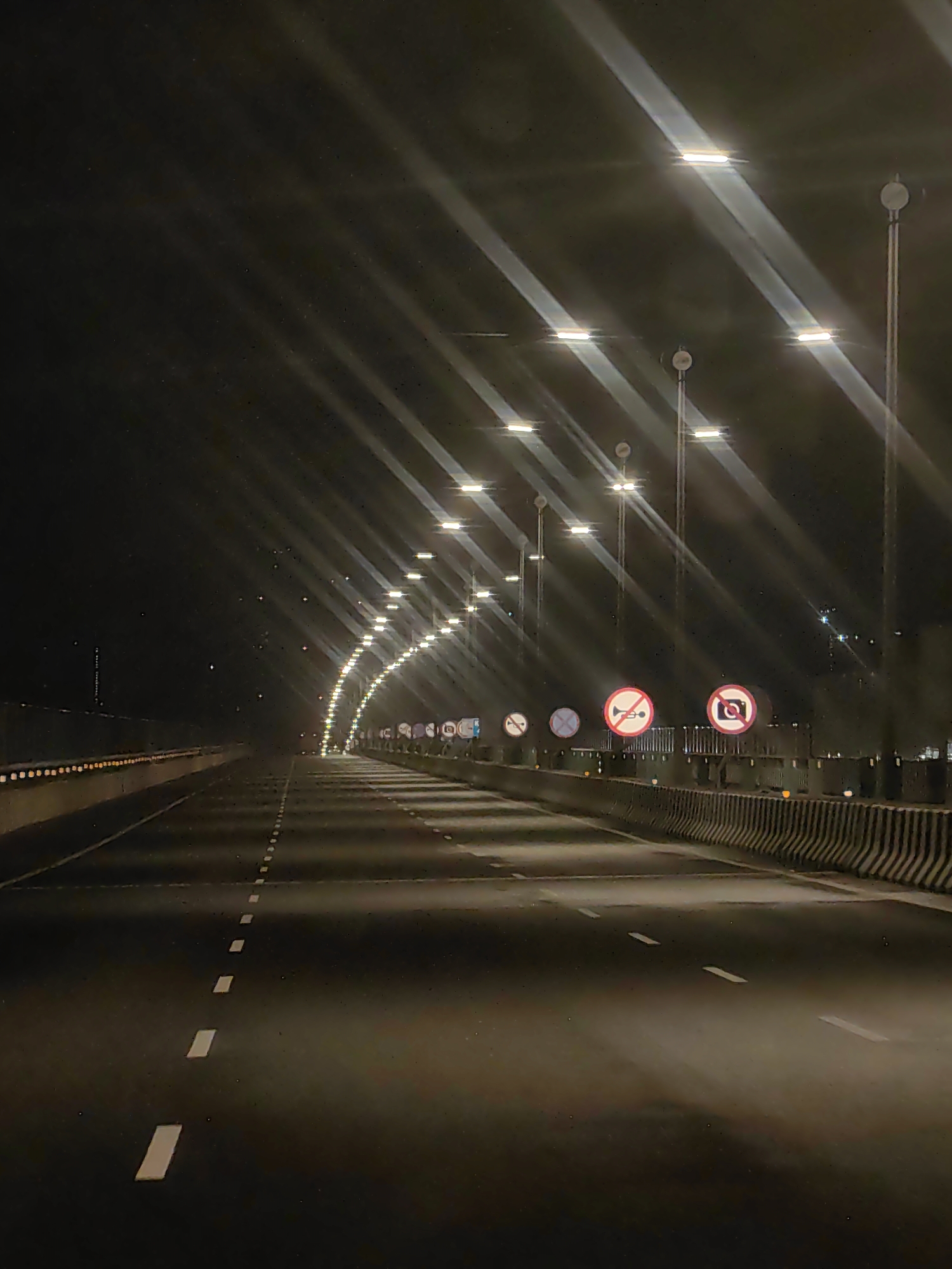 The image depicts a nighttime view of a highway in Nhava Khadi, India. The highway is illuminated by rows of streetlights mounted on poles, casting long, diagonal beams of light across the empty asphalt road. The road features white dashed lane markings. On the right side of the highway, there is a barrier with several circular traffic signs. One sign depicts a horn with a diagonal line through it, indicating no honking. Another sign shows a camera with a diagonal line through it, signifying no photography. The environment is dark, with the primary light sources being the streetlights and the headlights of unseen vehicles, creating a sense of depth and highlighting the road's curvature. The scene appears to be on an elevated road or bridge, with a concrete barrier lining the right side.