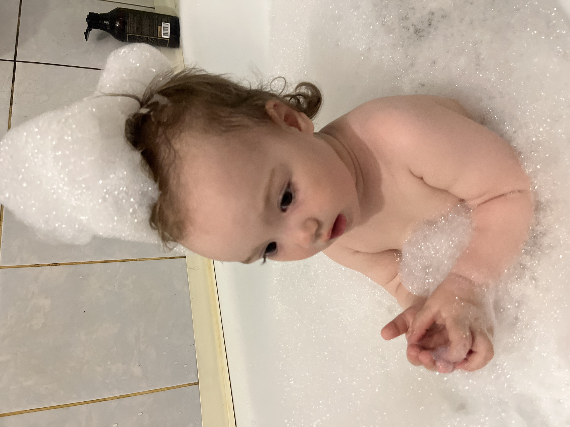 A baby is in a bathtub filled with bubbles. Some bubbles are on the baby's head like a hat. There is a bottle of dark-colored liquid on the tiled edge of the tub. The location is not identifiable as Shushary, Russia, from the provided image.