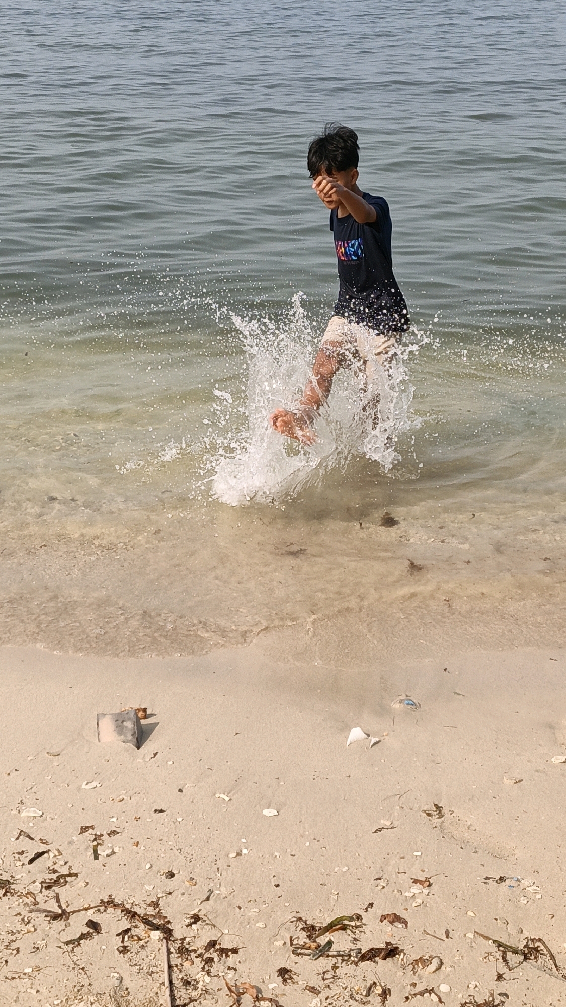A young boy with dark hair is captured in mid-air as he runs through shallow water at a beach in Cikal, Indonesia. He is wearing a dark t-shirt with a colorful graphic and light-colored shorts. Water splashes around his legs and feet, creating a dynamic effect. The background consists of the gently rippling surface of the ocean. The foreground shows a sandy beach with scattered shells and dried seaweed. The lighting suggests it is daytime.