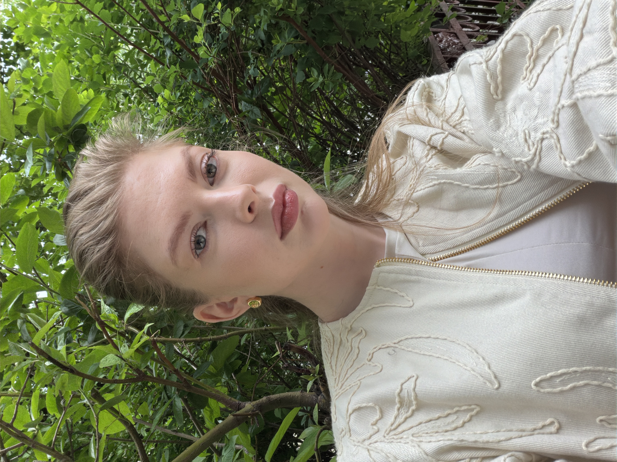 A woman is posing for a selfie outdoors, surrounded by lush green foliage. She has light blonde hair, fair skin, and is wearing a white jacket with embroidered details and a matching top. She is wearing a gold earring. The location appears to be a park or garden, and due to the abundant greenery, it's difficult to pinpoint the exact location within Moscow. The scene is well-lit, suggesting it's daytime.