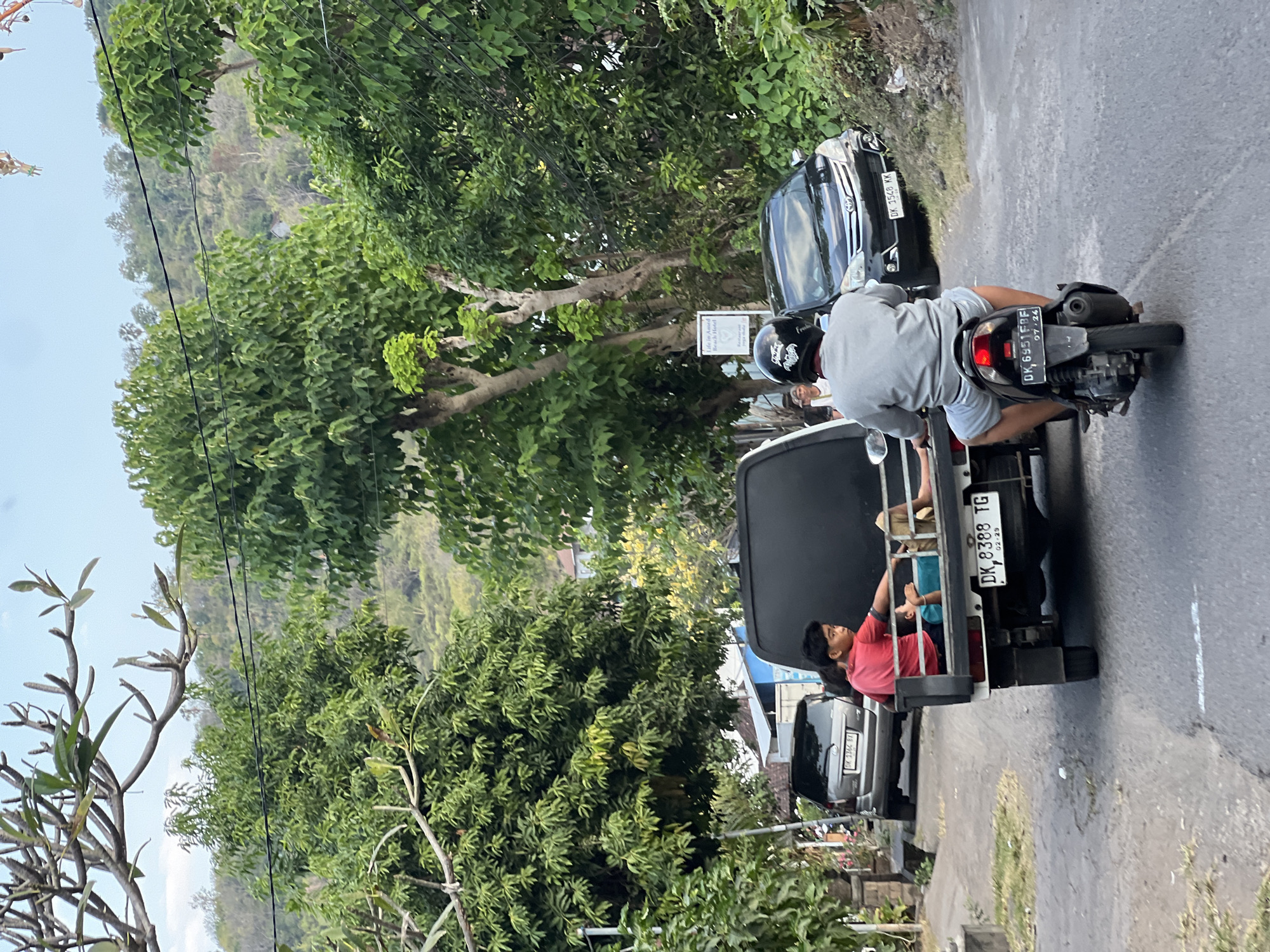 The image depicts a street scene in Abang, Indonesia. In the foreground, a person wearing a gray shirt and shorts rides a motorcycle, facing away from the camera. Behind them, a white pickup truck is visible, with two young individuals in the truck bed. One, wearing a red shirt, is seated, while the other, in a turquoise shirt, is lying down. A silver car is parked on the left side of the road, and a black car is in motion on the right. Lush green trees and foliage line the street, with a hilly, wooded area in the background. Overhead power lines are also present. The road appears to be paved, with some vegetation growing along its edges. The sky is overcast.