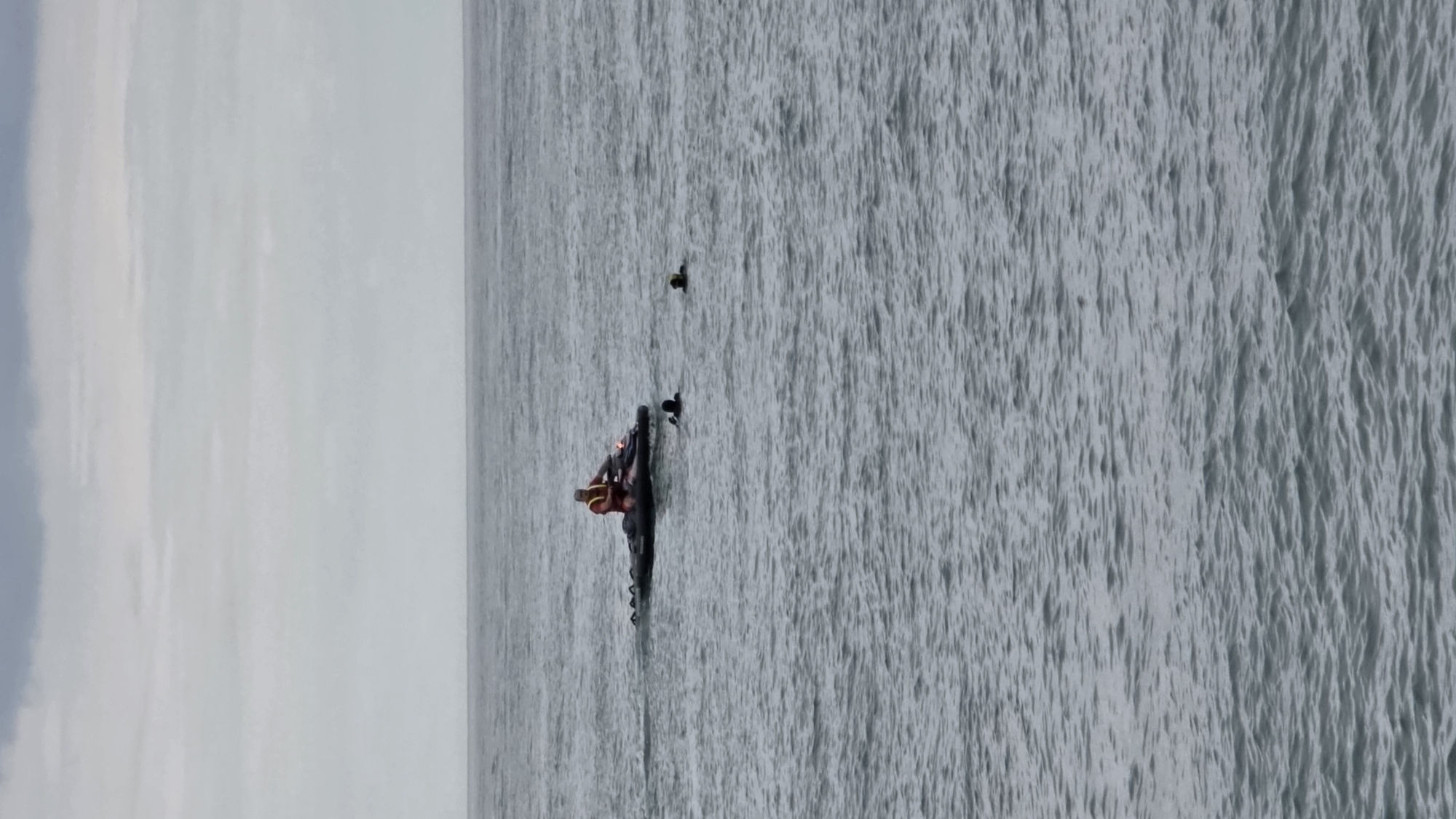 A jet ski, ridden by a person wearing a bright orange life vest, navigates choppy grey water. Two individuals, wearing dark attire and caps, are in the water near the jet ski, appearing to be swimming or treading water. A third person in dark attire is also in the water further away. The sky is overcast and grey, suggesting a cloudy day. The scene is consistent with an incident or activity taking place in the Black Sea off the coast of Batumi, Georgia, possibly a rescue operation or a water sport.