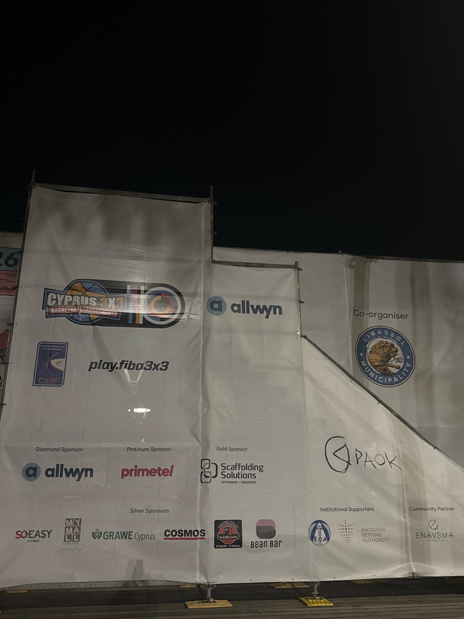 This is a nighttime, eye-level, medium shot of a white banner displaying various logos and text. The banner appears to be part of an outdoor event, possibly related to sports or a festival, as suggested by the "CYPRUS 3x3 BASKETBALL TOURNAMENT" logo. The banner is illuminated from the front, with some shadows visible.
The banner is divided into sections indicating different sponsorship tiers: "Diamond Sponsor," "Platinum Sponsor," "Gold Sponsor," "Silver Sponsors," "Institutional Supporters," and "Community Partner."
Under "Diamond Sponsor," the logo for "allwyn" is visible.
Under "Platinum Sponsor," the logo for "primetel" is visible.
Under "Gold Sponsor," the logo for "Scaffolding Solutions" is visible.
Under "Silver Sponsors," logos for "SO EASY STORES," "MY MA LL LIMASSOL," "GRAWEE Cyprus," and "COSMOS" are present.
Under "Institutional Supporters," the "Limassol Municipality" logo is displayed, along with a logo for the "NATIONAL BETTING AUTHORITY."
Under "Community Partner," the "ENAVSMA FOUNDATION" logo is visible.
Additionally, there is a smaller logo with "CBF" and a shield, likely representing the Cyprus Basketball Federation. The "play.fiba3x3" website is also promoted on the banner.
In the upper right portion of the banner, "Co-organiser" is written above the "Limassol Municipality" logo, indicating that the municipality is a co-organizer of the event. The presence of the Limassol Municipality logo strongly suggests the event is taking place in the city of Limassol, Cyprus. A handwritten scribble of "CPAOK" is also visible in the upper right section.
The banner is supported by a metal frame and a wooden barrier is visible at the bottom, suggesting the event is held outdoors, possibly in a public space or a venue within Limassol. The sky above is dark, indicating it is nighttime.