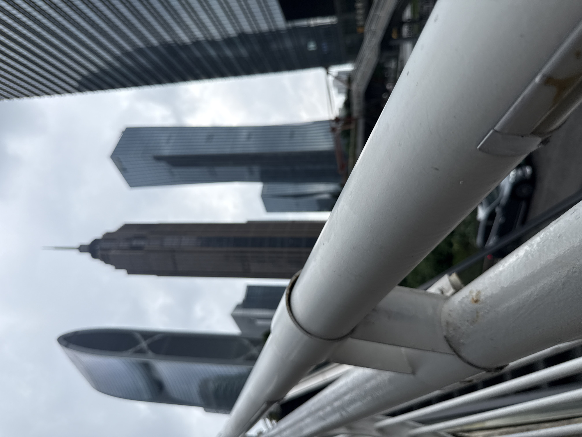 The image shows a perspective shot of several modern skyscrapers against a cloudy sky. In the foreground, a white metal railing or pipe is prominently featured, obscuring parts of the view. The railing is angled, suggesting the photo was taken from a higher vantage point, possibly from a bridge, elevated walkway, or another building.
The skyscrapers in the background display a variety of architectural styles. One building, in the middle distance, has a distinctive tiered or stepped design, culminating in a sharp spire. To its left, another skyscraper is more angular and geometric, appearing to be clad in glass. Further back and to the top left, a building with a striped facade is visible. The overall scene is urban and modern.
The weather appears to be overcast, indicated by the uniformly grey sky. There are no people visible in the frame. The presence of multiple tall buildings and the modern architecture suggest a bustling urban environment. The lighting is diffused, consistent with a cloudy day, and no specific time of day can be determined. The location is identified as Tianhe District, China. There is no visible text in the image. The composition, with the railing in the foreground, gives a sense of depth and perspective.